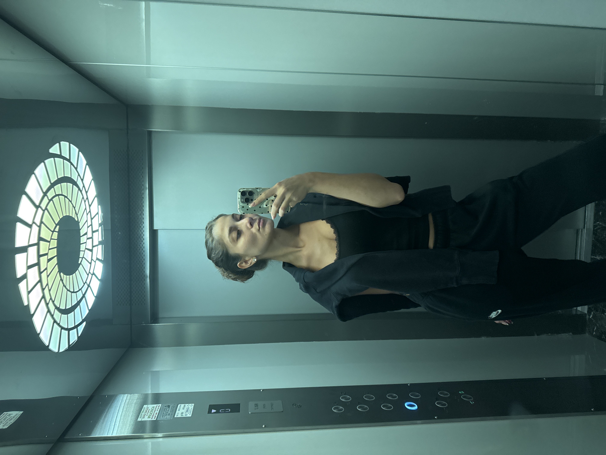 A woman is taking a selfie in an elevator. The elevator has a circular light fixture, a control panel with buttons, and signage in Bulgarian and Cyrillic alphabet. The location is likely in the city of Burgas, Bulgaria, given the language of the signage. The woman is wearing black clothing.