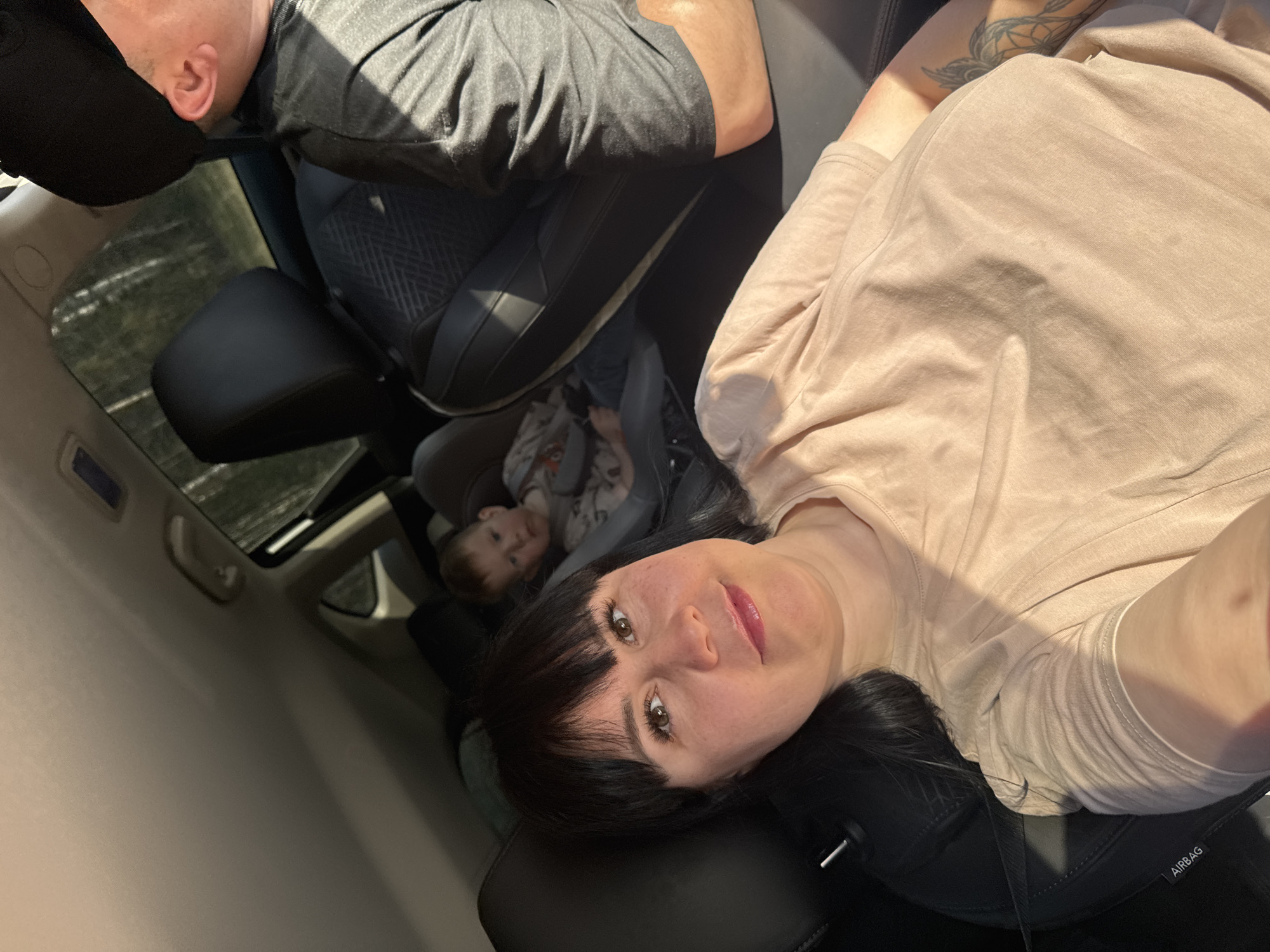 The image depicts the interior of a car, likely a selfie taken by a woman with dark hair and bangs. She is wearing a light-colored shirt. To the left of the woman is a child in a car seat, partially visible. Above them, the top of another person's head is visible, wearing a dark-colored cap and a dark-colored shirt. The car seems to have a sunroof. The location in Semenovo, Russia, cannot be confirmed from the image alone.