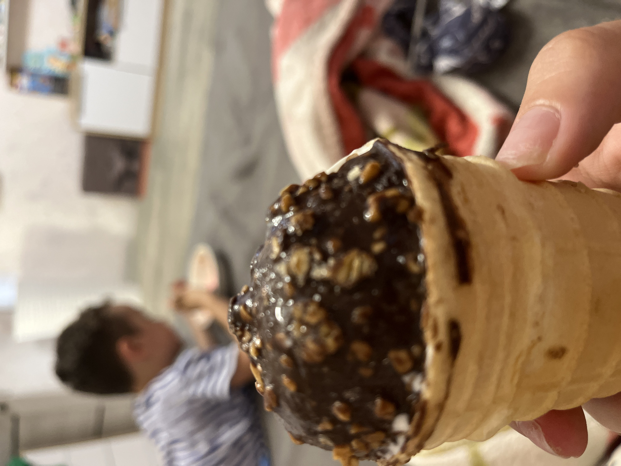 A close-up shot of a chocolate-dipped ice cream cone topped with nuts is held in a person's hand. In the blurred background, a young boy wearing a striped shirt is seated on the floor, facing away from the camera and looking towards a white bowl. The environment appears to be indoors, possibly a living room, with shelves and furniture visible in the distance. There is no specific indication of the city of Krasnyye Barrikady, Russia.