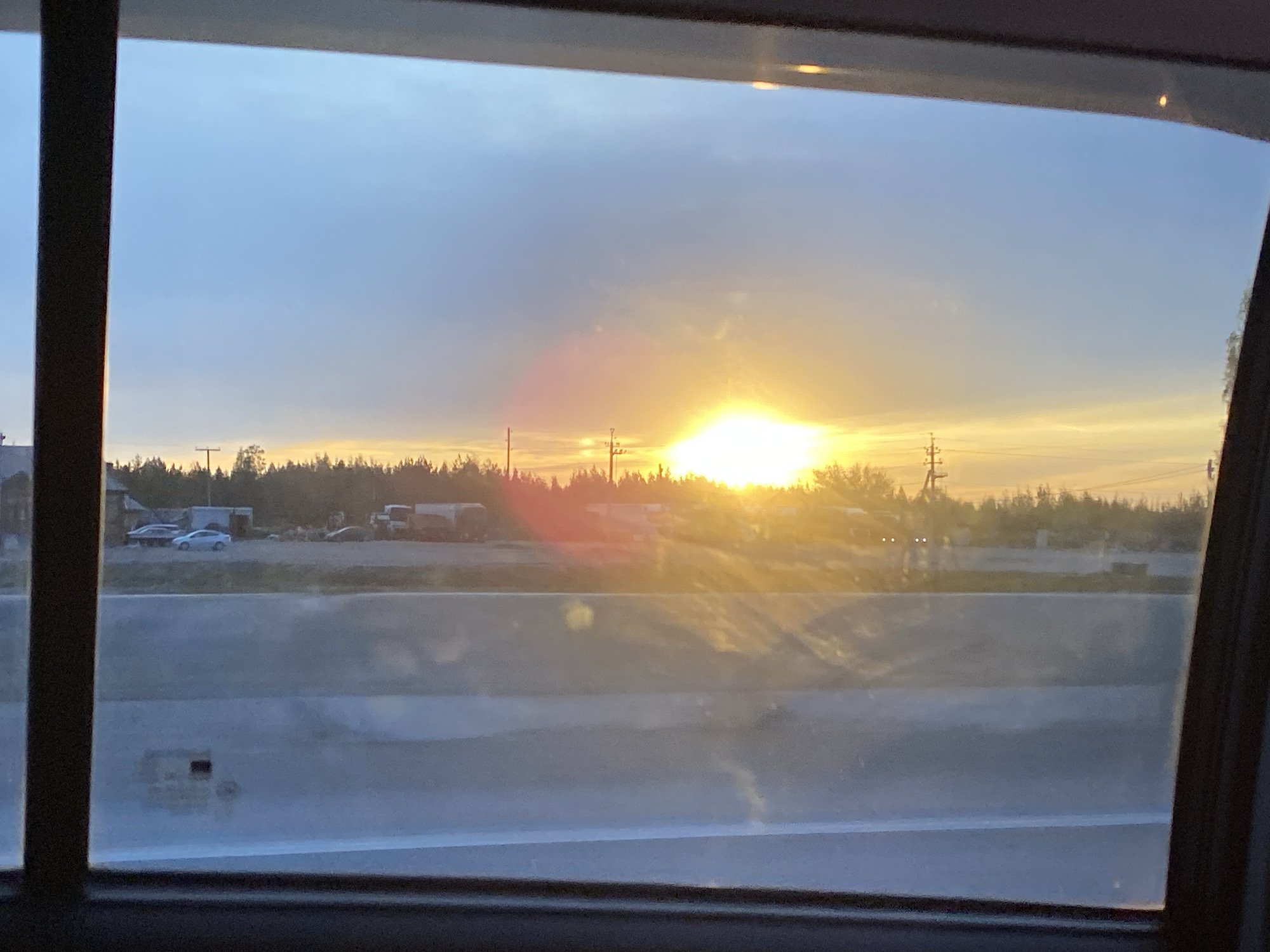 View from a car window with the setting sun visible over a highway in Bol'shiye Klyuchishchi, Russia. A forest and utility poles line the horizon behind the highway, and some parked vehicles are visible to the left. The highway barrier is in the foreground.
