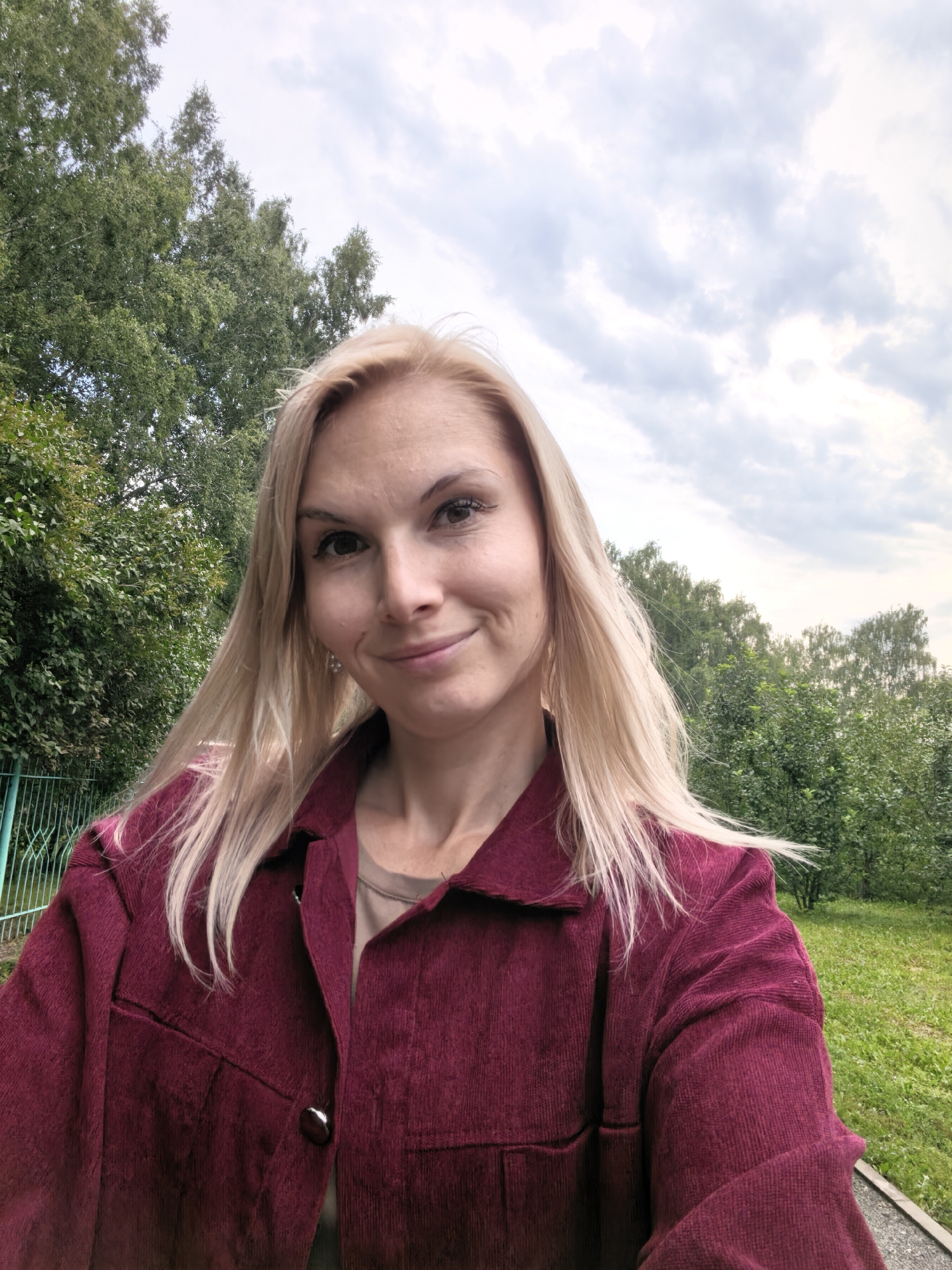 In Sterlitamak, Russia, a blonde woman with fair skin and light brown eyes is captured in a close-up, selfie-style photograph. She is wearing a maroon corduroy jacket over a beige shirt. Her hair is long and light blonde, falling loosely around her shoulders. The background consists of lush green trees and a cloudy sky, suggesting an outdoor setting, possibly a park or a residential area. A hint of a green metal fence is visible on the left side of the frame. The image appears to be taken during the daytime.
