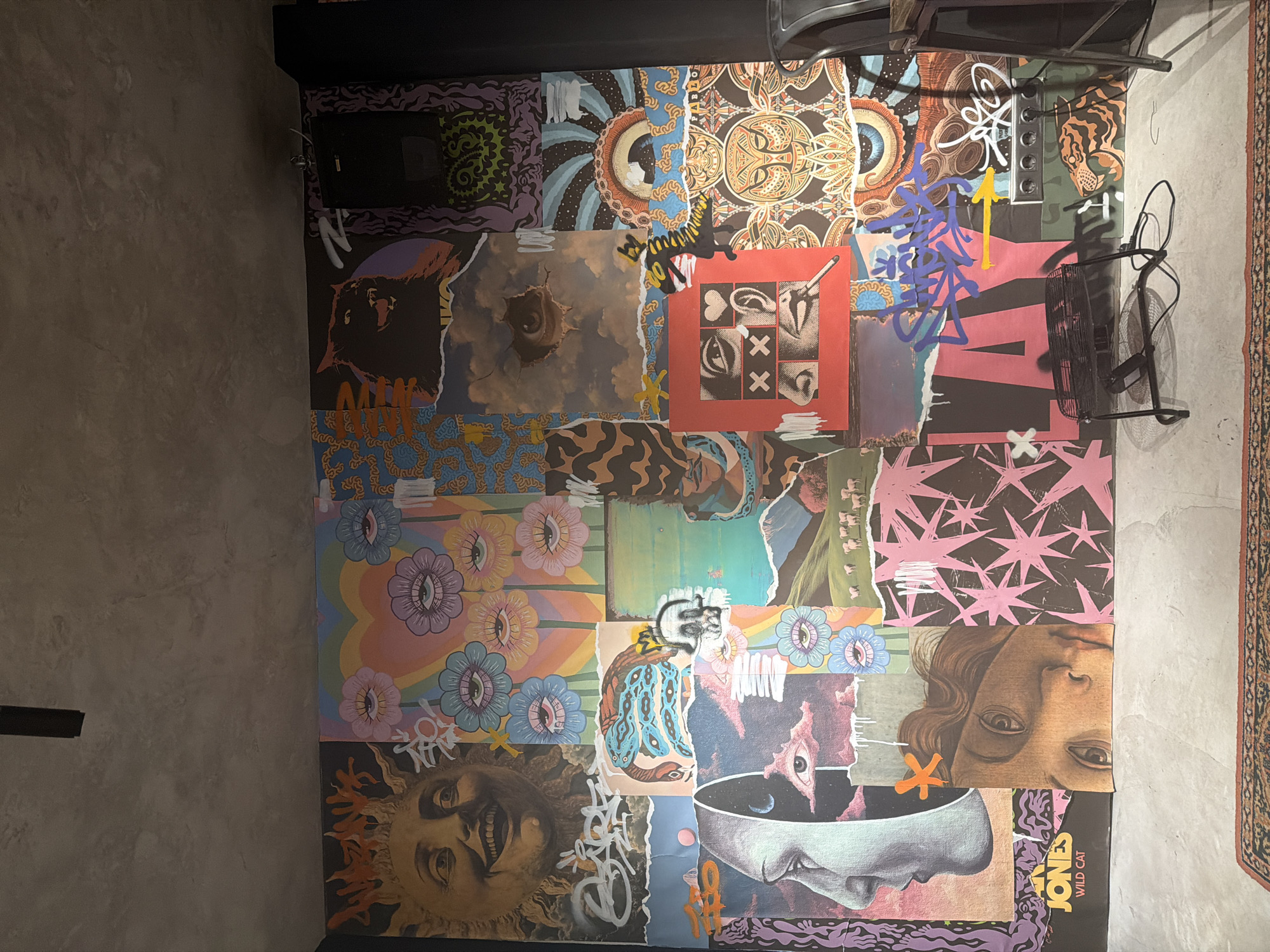 The image shows a collage of album covers and other graphic elements arranged on a flat surface, possibly a floor. The collage is composed of various imagery, including faces, eyes, abstract patterns, and graffiti-like markings. A black metal side table with a wire basket sits on top of the collage. The environment appears to be a room with a gray concrete wall to the left and a rug partially visible on the right. The location is unknown.