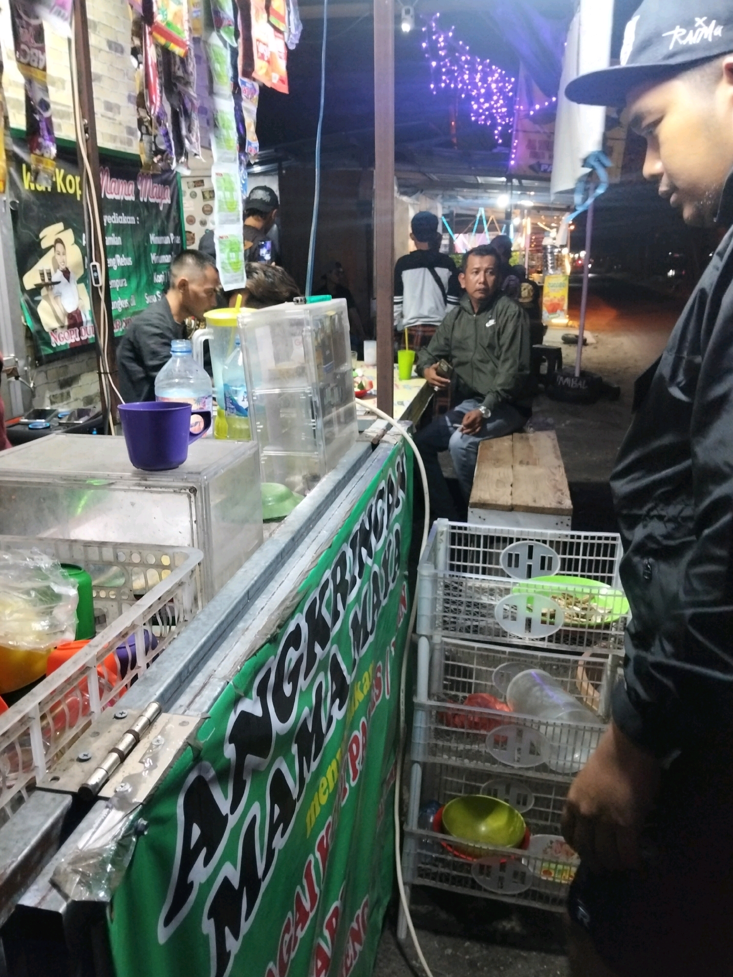 A close-up, eye-level shot captures a street food stall at night in Denpasar, Indonesia. The stall features a green banner with white text, partially visible and appearing to advertise food or drinks. To the left, a clear plastic container displays stacked dishes. In the center, a purple mug sits on a clear plastic surface. Behind the counter, several individuals are present. A man wearing a dark jacket and a cap is visible in the foreground, facing right, with his profile captured. Further back, another man in a green jacket and jeans is seated on a wooden bench, smoking a cigarette. A stack of white plastic shelves holding various items, including bowls and plates, is positioned to the right of the bench. The background is blurred, showing dimly lit stalls and what appears to be decorative lighting. The scene suggests a bustling night market or roadside eatery.