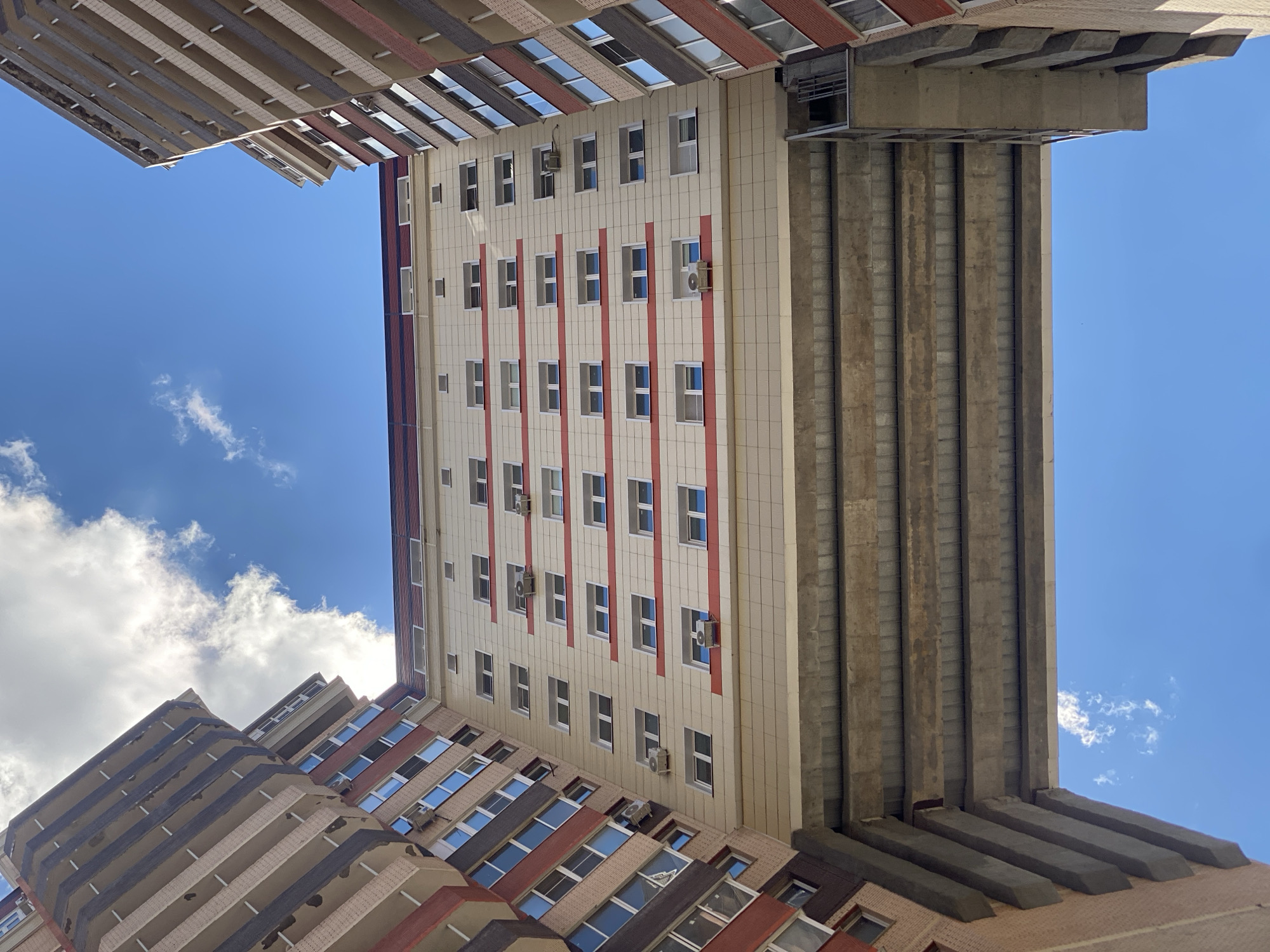 This media file shows a low-angle, upward view of multiple apartment buildings and a concrete structure against a bright blue sky with scattered clouds. The buildings are modern, with many windows and balconies. One building is cream-colored with prominent red vertical stripes. Another building, partially visible on the left, has a facade with alternating dark and light colored balconies. A large, imposing concrete structure, likely part of an overpass or elevated walkway, dominates the right side of the frame, casting shadows and creating a stark contrast with the buildings and sky.
There are no people visible in the image. The objects include the buildings themselves, windows, balconies, and air conditioning units mounted on the exterior walls of one building. The scene is an urban environment, characterized by residential high-rises.
The lighting suggests it is daytime, likely midday, given the bright sunlight and the shadows. The weather appears to be clear and sunny, with a few wispy clouds. There are no explicit location cues in terms of signs or landmarks, but the mention of Kotelniki, Russia, provides geographical context.
No visible text is present in the image. The overall impression is one of urban density and architectural scale, viewed from an unusual perspective that emphasizes the verticality and the expanse of the sky.