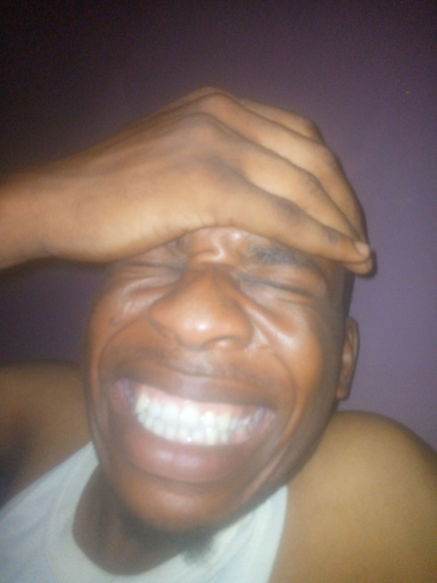 A close-up photograph shows a dark-skinned man with a hand on his forehead and his eyes closed. He is smiling widely, exposing his teeth. He is wearing a white tank top. The background appears to be a solid light purple color. No specific details identify the location as Ikorodu, Nigeria.