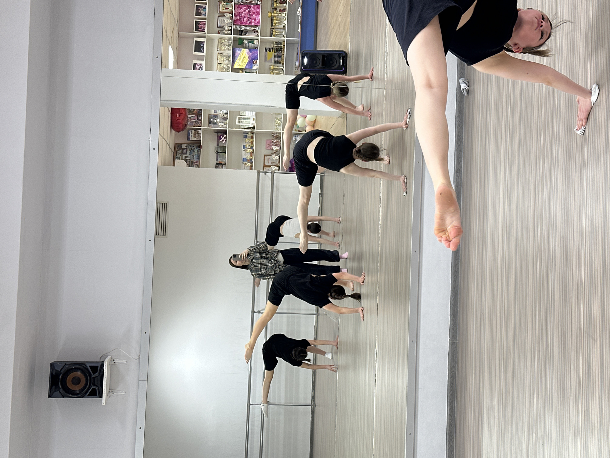 The image captures a dance or exercise studio, potentially in Votkinsk, Russia, as specified. Multiple individuals are engaged in a stretching or exercise routine, seemingly instructed by one person. The people are primarily women and girls, mostly dressed in black athletic wear. The studio includes a barre, likely used for balance and support during exercises. A large mirror reflects the scene, creating a sense of depth. In the background, shelves display trophies, awards, and various objects. A black speaker is mounted on a wall. The flooring is a light, patterned material. Overall, the setting is a dance or exercise studio where a group is practicing.