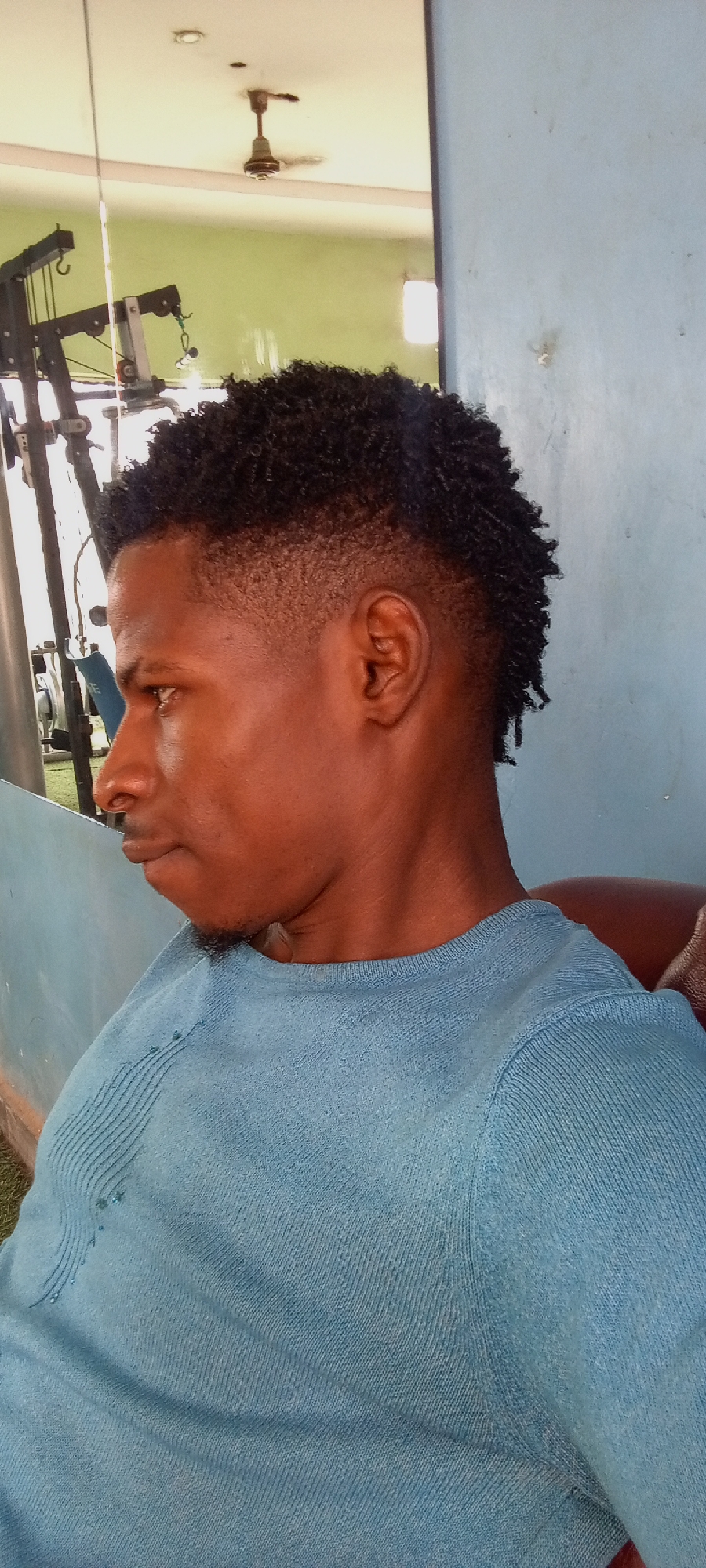 A medium shot presents a dark-skinned man in a light blue sweater, seated in a room that appears to be a gym, likely in Akwanga, Nigeria. The man is captured in profile, facing left, with a distinct haircut featuring short, tightly curled hair on top that gradually tapers down on the sides. He has a small amount of facial hair around his chin.
Behind him, gym equipment is visible, reflected in a mirror. There is a ceiling fan, and a wall painted blue.