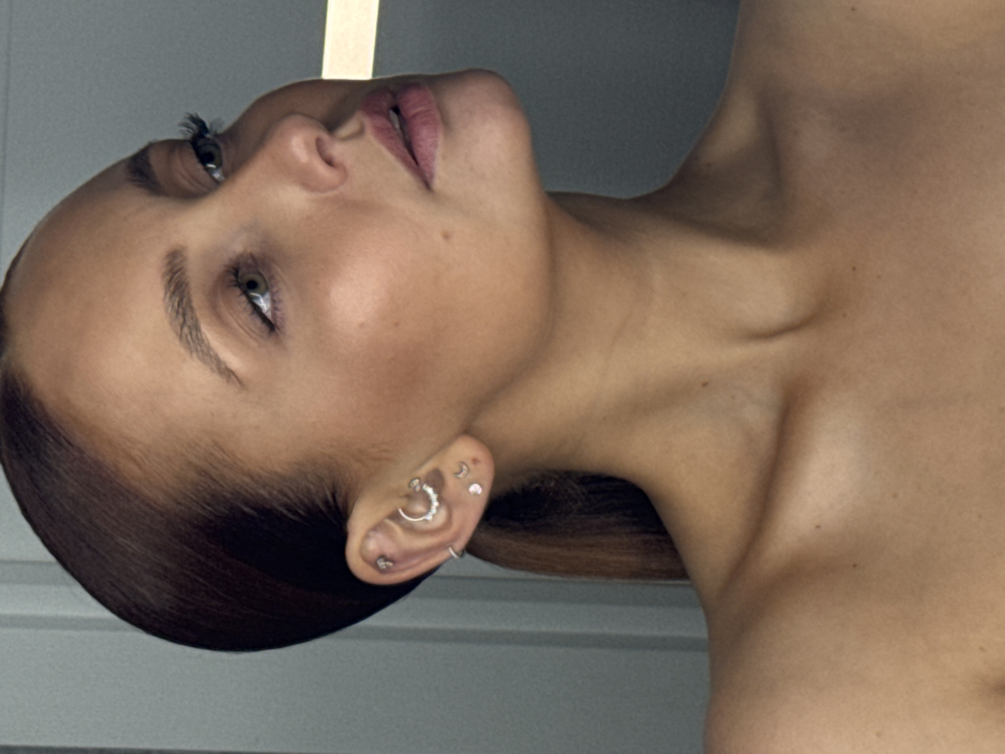 A medium close-up portrait features a woman with her head tilted significantly back and to her left, with the image rotated counter-clockwise, presenting her face almost horizontally. Her gaze is directed upwards and slightly to the left, conveying a serene or contemplative expression. She has clear, fair skin, a defined jawline, and smooth dark brown hair that appears pulled back. Her eyes are a striking greenish-brown, and her lips are subtly colored with a soft pink.
A prominent detail is her left ear, which is adorned with multiple silver-toned piercings: a helix piercing with a decorative curved barbell of small stones, a conch piercing with a small stud, two flat piercings featuring delicate crescent moon studs, and a simple hoop in her earlobe. Her bare shoulders and collarbone are visible, suggesting either nudity or a strapless garment.
The background consists of a plain, light gray wall with a vertical strip of bright white light visible on the far side of her head, possibly a light fixture or architectural element. The lighting is soft and diffused, emphasizing the natural contours of her face and upper body. No specific activity is depicted other than her pose. There is no visible text in the image. The media file itself contains no visual cues to suggest the provided location context of Chaikovsky, Russia.