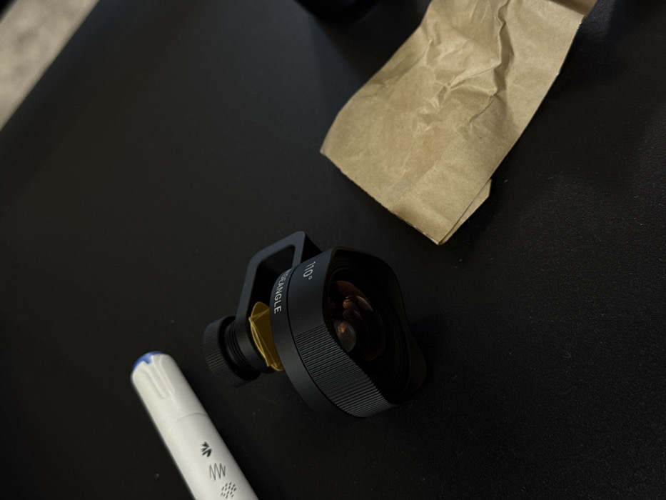 The scene is located in Moscow, Russia. It depicts a black, cylindrical wide-angle camera lens attachment, marked "110° WIDEANGLE," resting on a dark, matte surface. A gold-colored component is visible near the lens's mounting point. To the upper left, a white marker pen with a blue cap and black stylized markings on its barrel lies alongside the lens. To the lower right, a crumpled piece of light brown paper is present. No individuals or active interactions are depicted; the objects are stationary on the dark surface.