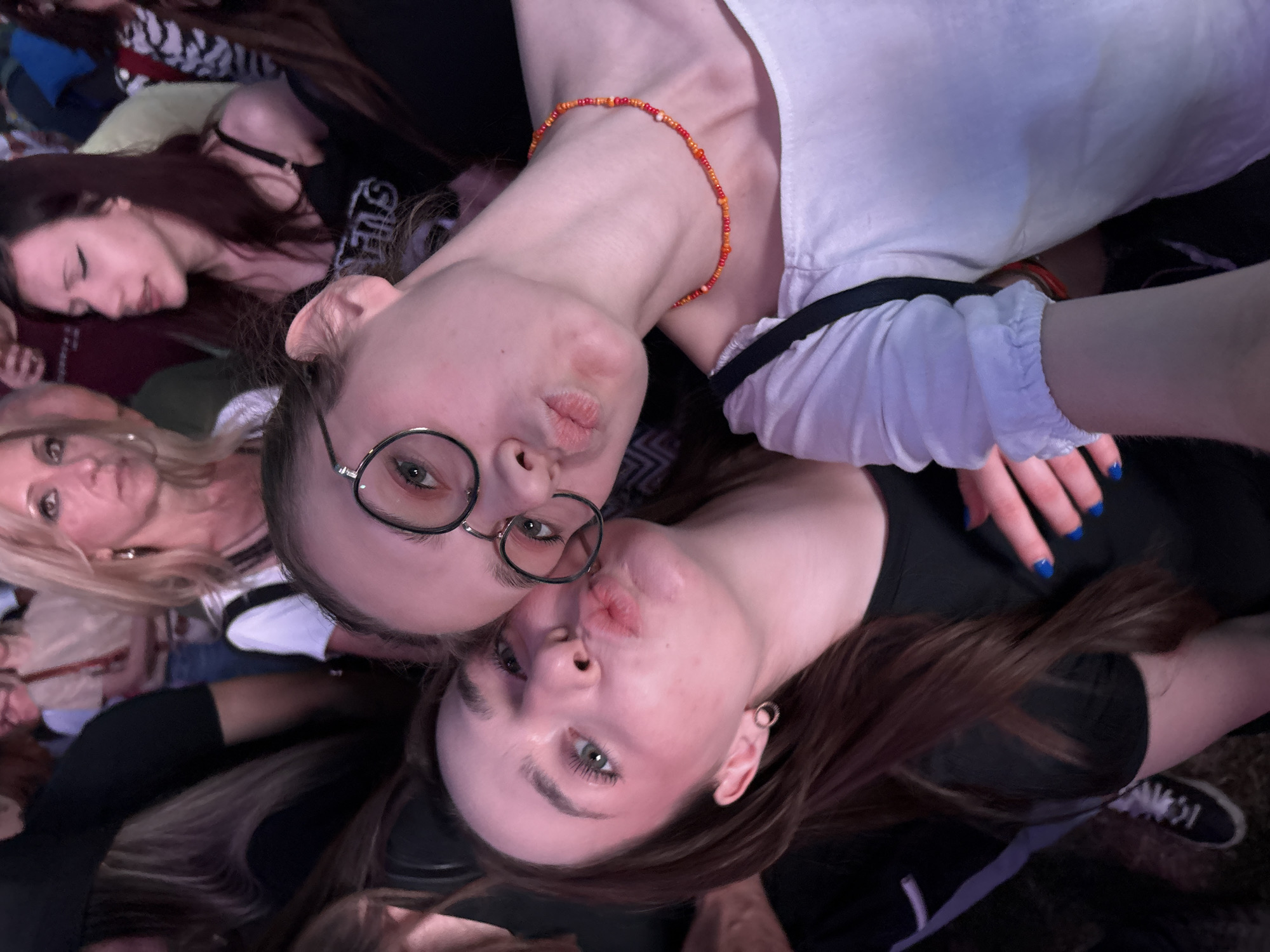 The image is a selfie taken from above, showing two young women puckering their lips as if to kiss. The woman on the left wears round glasses and a white top. She has a beaded orange and red necklace. The woman on the right has long brown hair and wears a black top. Her nails are painted blue. There are several other people in the background, including a blonde woman and a woman with dark hair lying down. The location of this image in Kragujevac, Serbia is unknown and not determinable from the image.