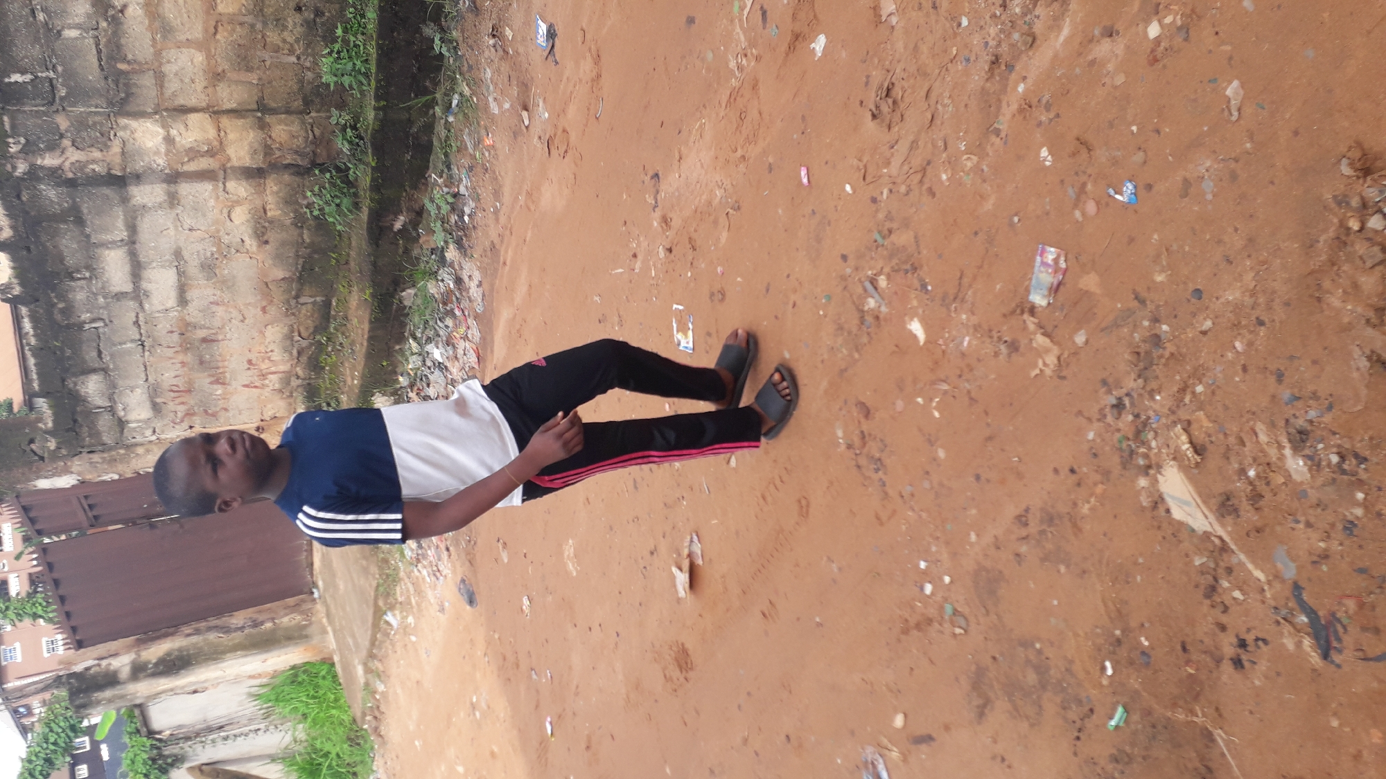 A young Black boy stands on a dirt road in Nkpor, Nigeria. He is wearing a blue and white t-shirt with white stripes on the sleeves and black pants with pink stripes down the sides. He is also wearing black slides on his feet. The road is unpaved and appears to be made of red dirt. There is a brick wall behind the boy, and a metal gate to the left. The area is littered with debris, including plastic bags and wrappers. The background shows some greenery and buildings. The boy is looking to his right and his body is angled to his left.