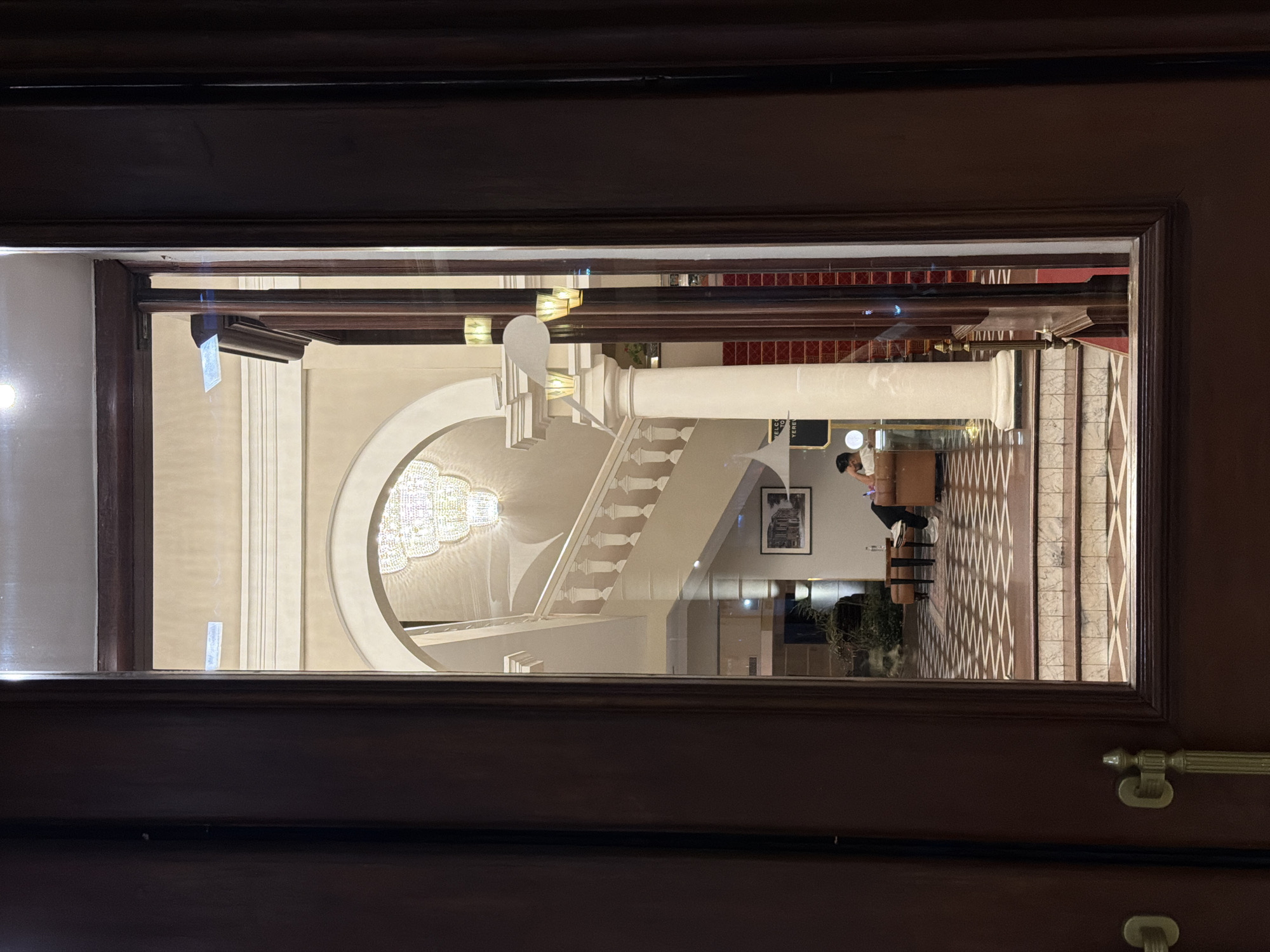 The original image is presented vertically, rotated 90 degrees clockwise. When viewed upright, it captures a glimpse into a grand and elegantly appointed interior space, likely a hotel lobby or a similar public building, framed through a dark wooden door or window panel.
The central features of the interior include a prominent white classical column and a sweeping grand staircase with a white balustrade, ascending towards an upper level. Overhead, a large, ornate multi-tiered crystal chandelier hangs, casting warm light and reflections throughout the area.
On the lower right side of the view, a person is seated at a dark wooden counter or desk, possibly engaged in work, attending to a guest, or waiting. They are wearing a light-colored top and dark pants. Behind them, a framed black-and-white picture adorns a wall.
The floor features intricate geometric tiling in shades of brown, white, and beige. A small black sign with white text is visible near the seated person, displaying "WELCOME TO YEREVAN". The dark wooden door frame and a brass handle are visible in the foreground, suggesting the viewer is looking into the space from an adjacent room or hallway. The overall ambiance suggests a well-maintained, luxurious, and stately environment, illuminated primarily by artificial lights, indicating it could be evening or simply a naturally dimmer indoor setting in Yerevan, Armenia.