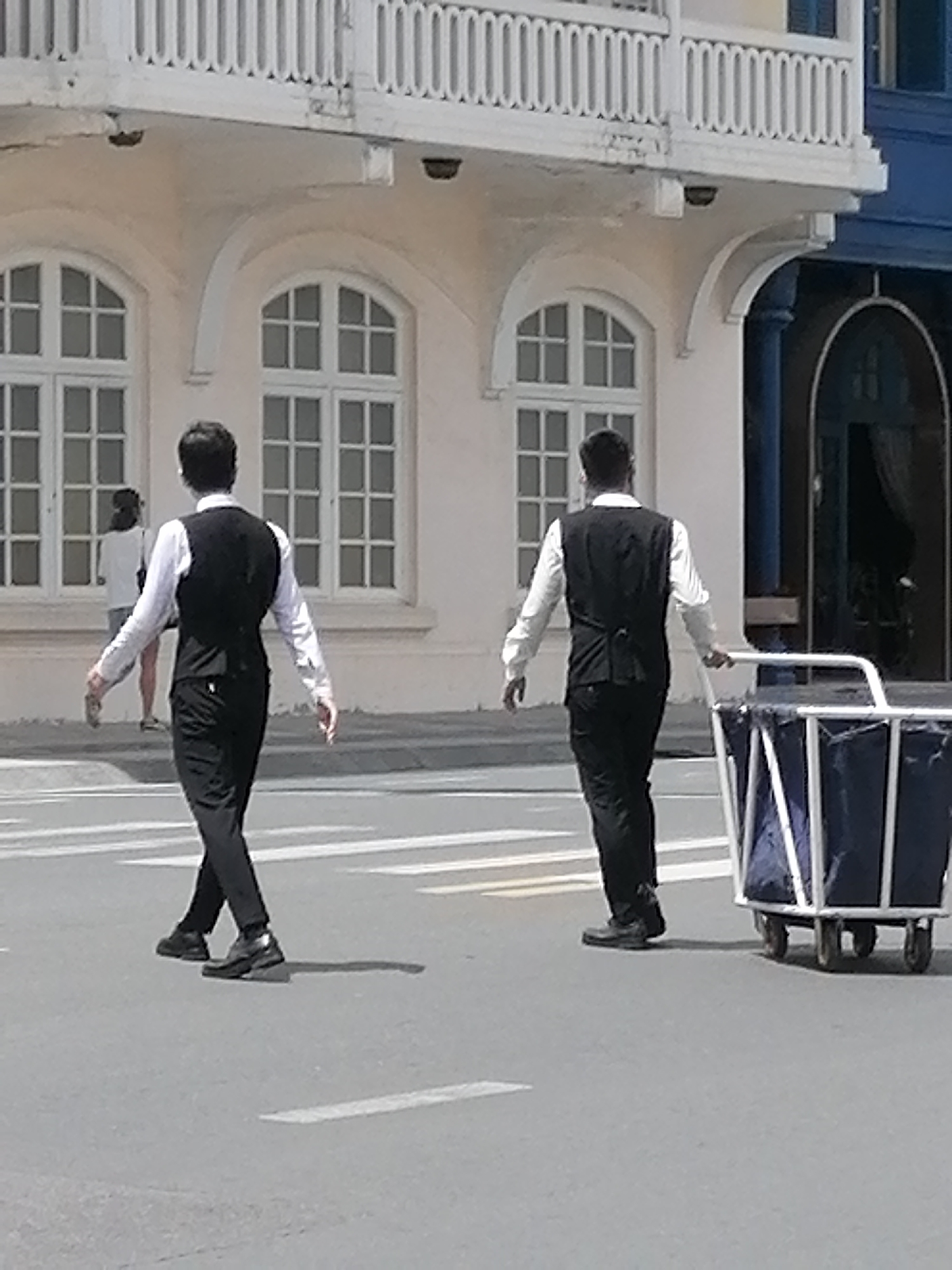 Two men in formal attire are walking across a crosswalk in front of a light-colored building. The man on the left is wearing a white dress shirt and a black vest, with black pants and black shoes. He is walking with his arms slightly out to his sides. The man on the right is also wearing a white dress shirt and a black vest with black pants and black shoes. He is pushing a white metal cart with a blue tarp covering its contents. The building behind them has multiple arched windows and a white railing on the upper level. The ground is asphalt with white painted crosswalk lines. The scene appears to be outdoors in a city setting. The presence of a building with colonial architectural features and the general ambiance suggest a location like Da Lat, Vietnam, a city known for its French colonial architecture and pleasant climate.