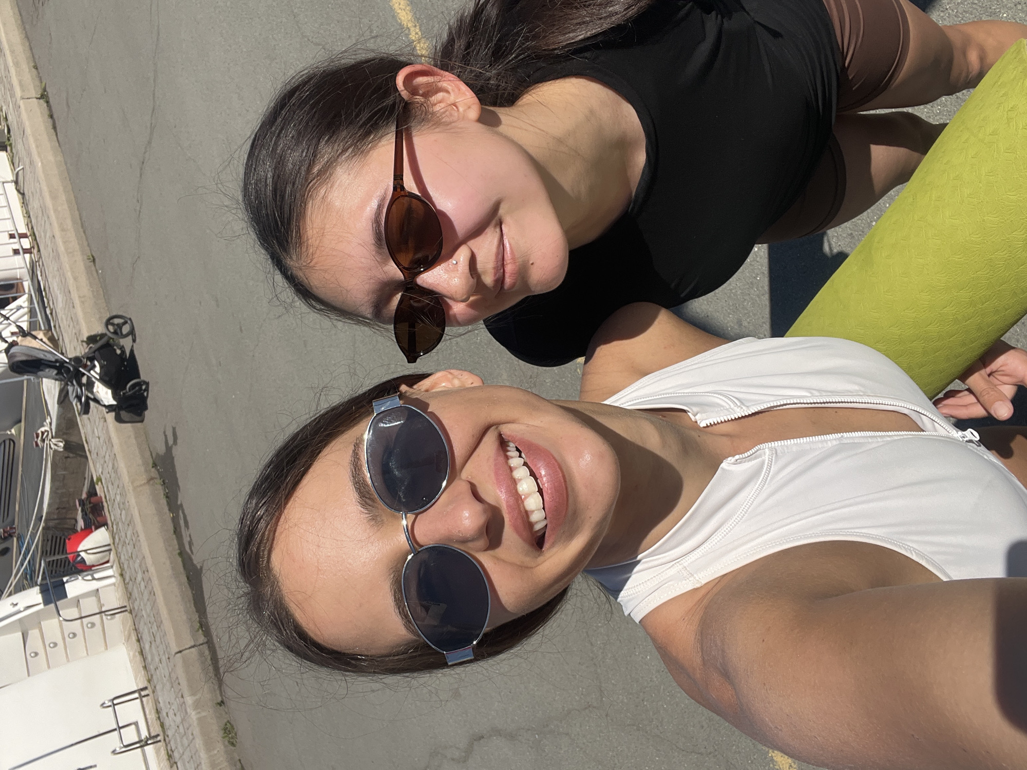 Two young women are posing for a selfie outdoors on a sunny day. The woman in the foreground is wearing a white athletic top with a zipper and silver framed sunglasses. She is smiling broadly, revealing her teeth. Behind her, the other woman is wearing a black top, brown shorts, and brown framed sunglasses. She is also smiling. A green yoga mat is visible near the women. The background shows a portion of a white yacht docked next to a stone curb and pavement. The photo was taken near the water in an urban setting. The location is likely Vladivostok, Russia, based on the marina context.