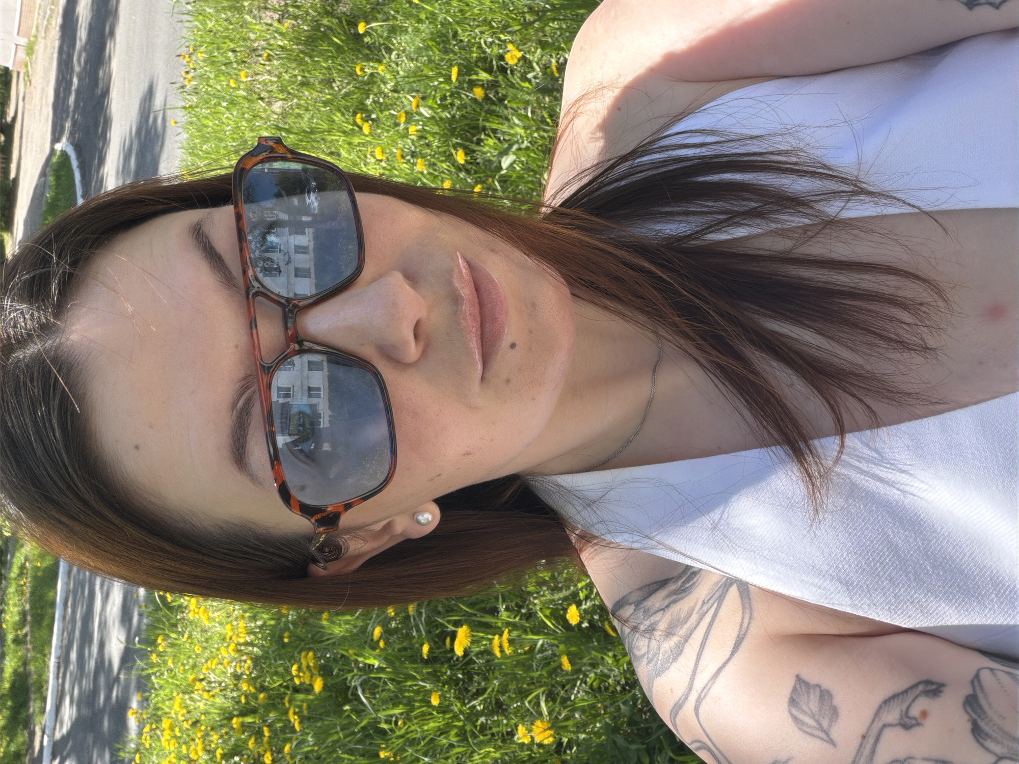A woman with brown hair and sunglasses poses in a green, grassy area dotted with yellow dandelions. Her tattoos are visible on her arm and the reflections in her sunglasses show a white building and blue sky. A paved road can be seen to the side. The image appears to be taken in a park or grassy area. The city location is Chaikovsky, Russia.