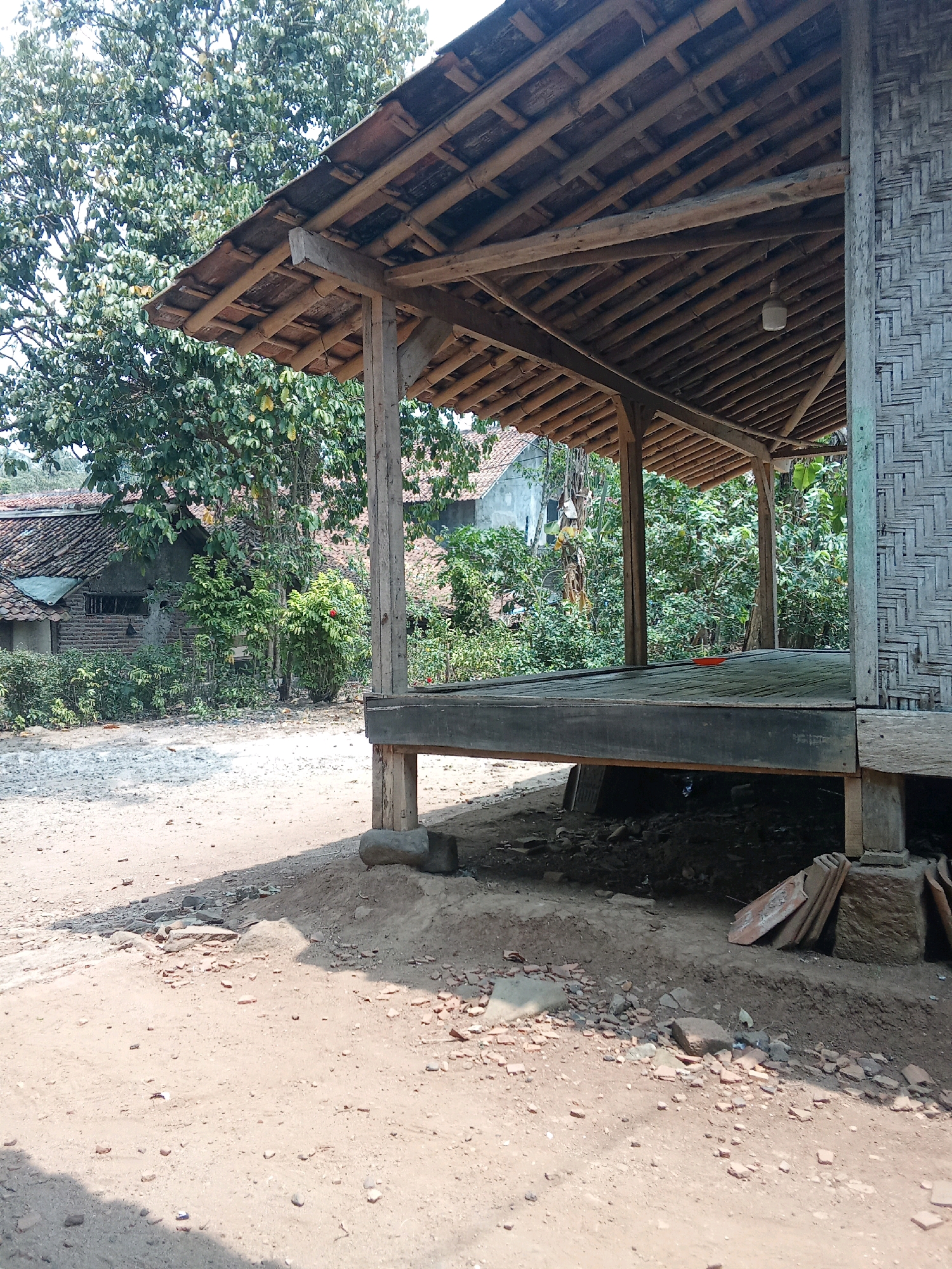 The image depicts an outdoor scene in Cempaka, Indonesia. In the foreground, a dirt path is visible with scattered debris, including small stones and broken pieces of reddish-brown material, possibly terracotta tiles. A shadow stretches across the path from the left. To the right, a raised wooden platform is supported by thick wooden posts. The platform has a weathered wooden floor. The side of the platform is constructed from woven bamboo. The roof of the structure is made of wooden beams and what appears to be terracotta tiles. In the background, various buildings with tiled roofs and walls made of brick or concrete are visible, partially obscured by lush green foliage. Tall trees with dense leaves are also present. The overall impression is one of a rural or semi-rural setting.