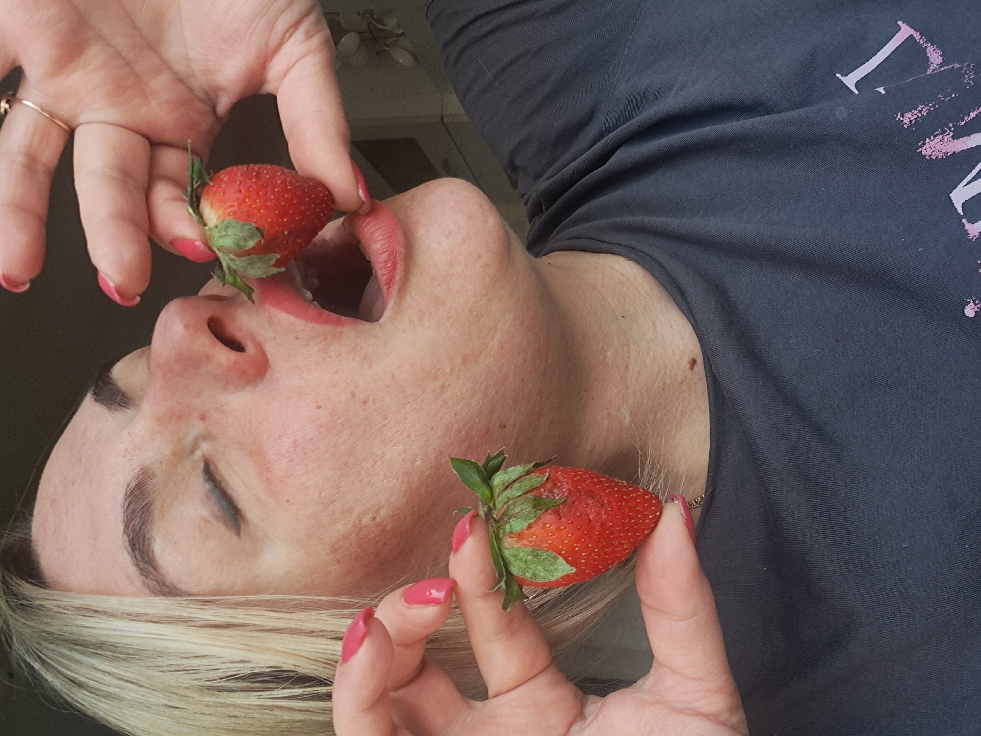 A fair-skinned, light-blonde-haired woman is holding two strawberries near her mouth. The woman has pink nail polish on her fingernails and is wearing a t-shirt with a faded pink and white graphic. Her mouth is open, and one strawberry is close to entering her mouth. The woman's eyes are closed. The setting appears to be indoors. The location of Elektrostal, Russia, is not discernible from the image.