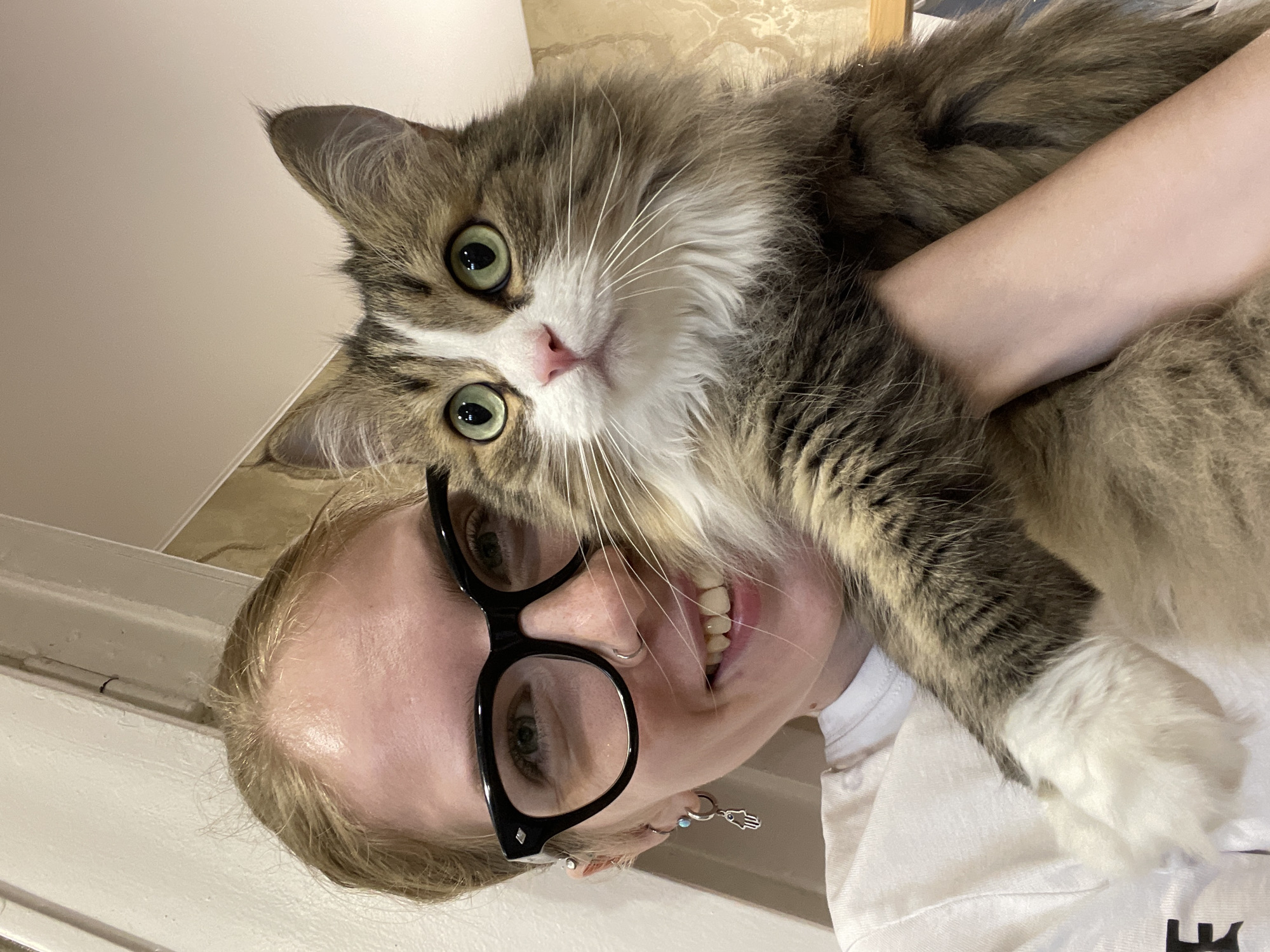 The image presents a close-up, rotated portrait of a person and a cat in an indoor setting. The person, who appears to be a woman, has light skin, light-colored hair, and wears large black-rimmed glasses. She is smiling broadly, showing her teeth, and has a nose piercing and an earring featuring a hamsa hand charm with a light blue stone. She is wearing a white t-shirt.
Cradled in the person's arms is a fluffy, long-haired cat with prominent green eyes, a pink nose, and long white whiskers. Its fur has a distinctive brown/grey tabby pattern mixed with white fur on its face, chest, and paws. The cat is looking directly at the camera with a calm and engaging expression.
The scene captures an affectionate interaction between the person and the cat, likely a selfie or a close-up photograph taken from an elevated angle. The background consists of simple, light-colored walls and possibly a textured surface, indicating a home environment. The lighting suggests indoor conditions. The overall emotion conveyed is one of warmth, happiness, and companionship. The image is rotated 90 degrees clockwise from its original capture orientation. There is no visible text within the image. The location context provided is Izhevsk, Russia.