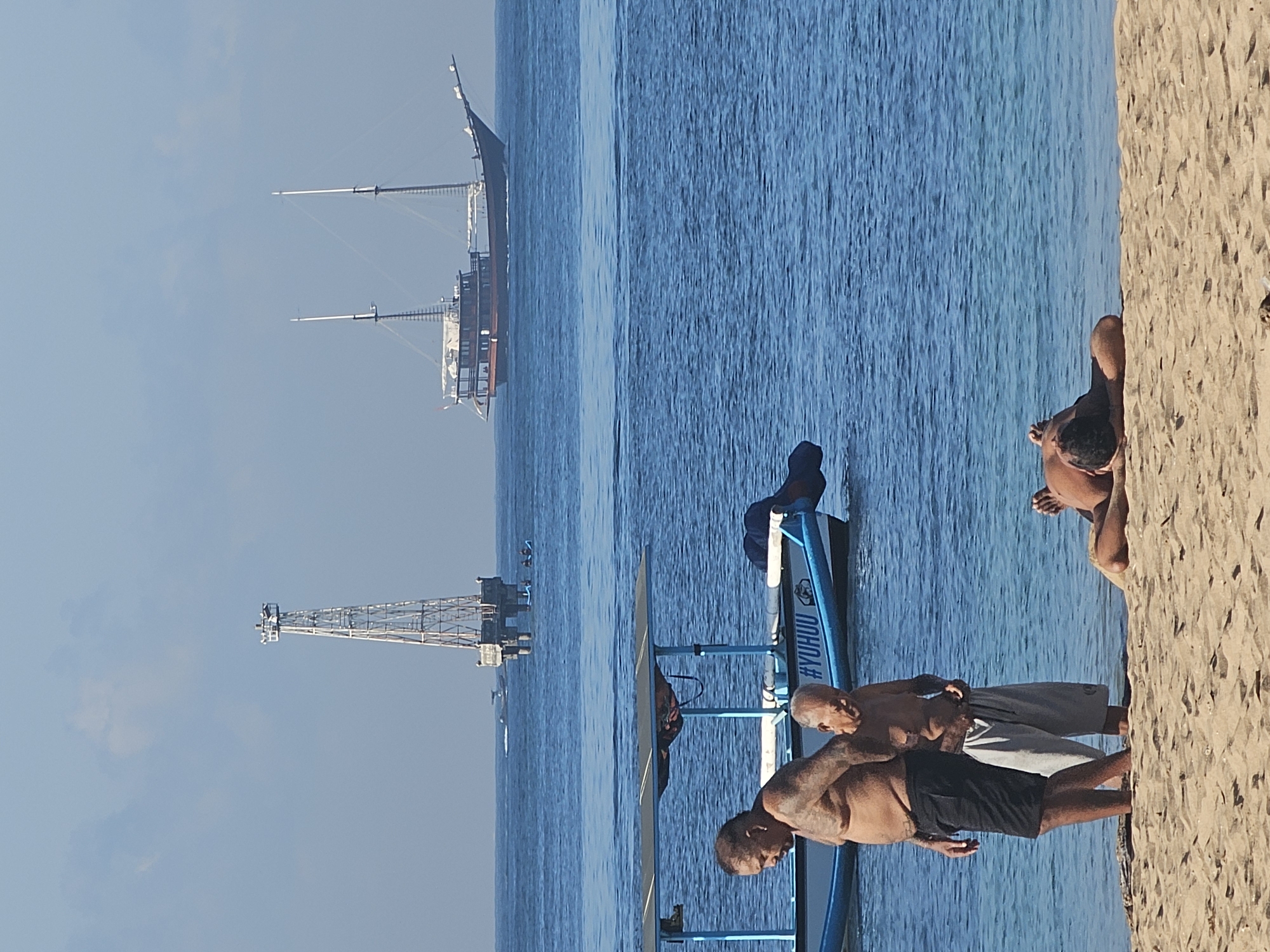 This image shows a beach scene in Denpasar, Indonesia. In the background, there's an offshore oil platform and a large sailing vessel on the water. Closer to the shore, a blue outrigger canoe is partially visible. In the foreground, on the sand, there are three shirtless men. One is standing near the canoe, wearing black shorts. Another is standing behind him, wearing grey shorts. A third man is lying face down on the beach. The water is clear and blue.