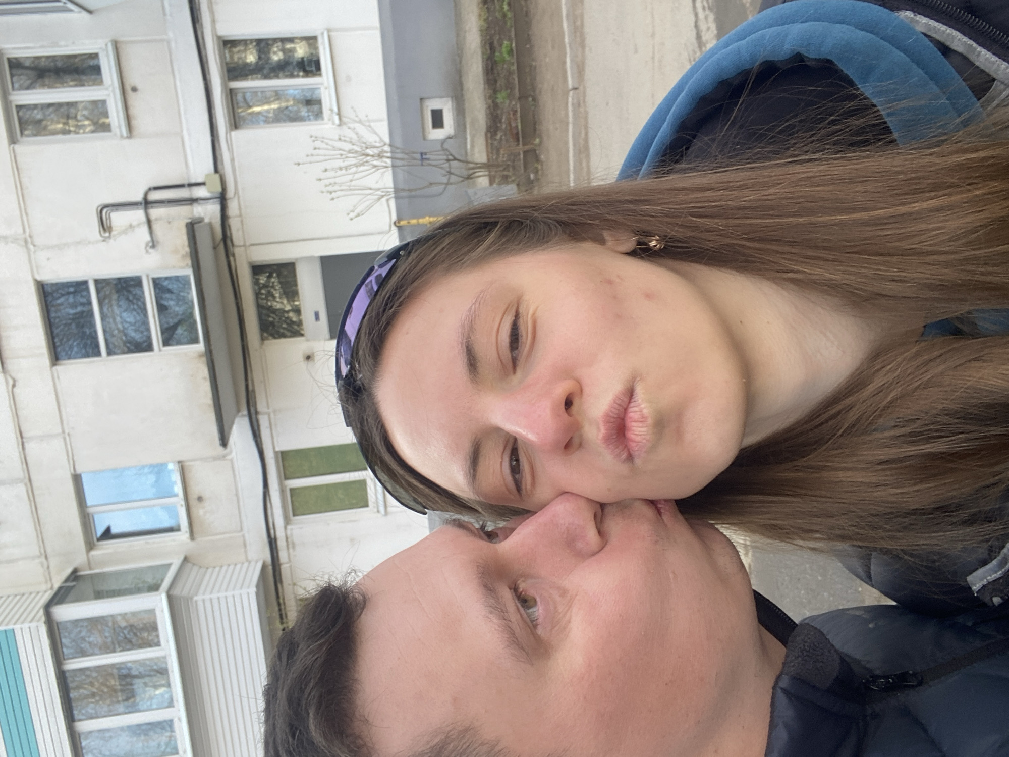A close-up, selfie-style photo captures a moment of affection between two young adults. The image is rotated counter-clockwise. A female subject, with long brown hair, a purple and black headband, and a blue hooded jacket, is on the right side of the frame, facing left. She has light eyes and is making a pouting face, bringing her lips close to the other person. To her left, partially obscured by her, is a male subject, visible from his nose to his forehead. His face is turned towards her, with his eye open and looking. His dark jacket is visible at the bottom of the frame.
The background shows a light-colored, multi-story building, likely an apartment block, with numerous windows reflecting bare trees or the sky. Some external pipes or conduits are visible on the building's facade. The overall setting appears to be outdoors, on a street or sidewalk adjacent to the building. The lighting suggests it is daytime, possibly late winter or early spring given the bare branches reflected. No visible text is present in the image. The scene conveys intimacy and affection. The location is Chaikovsky, Russia.