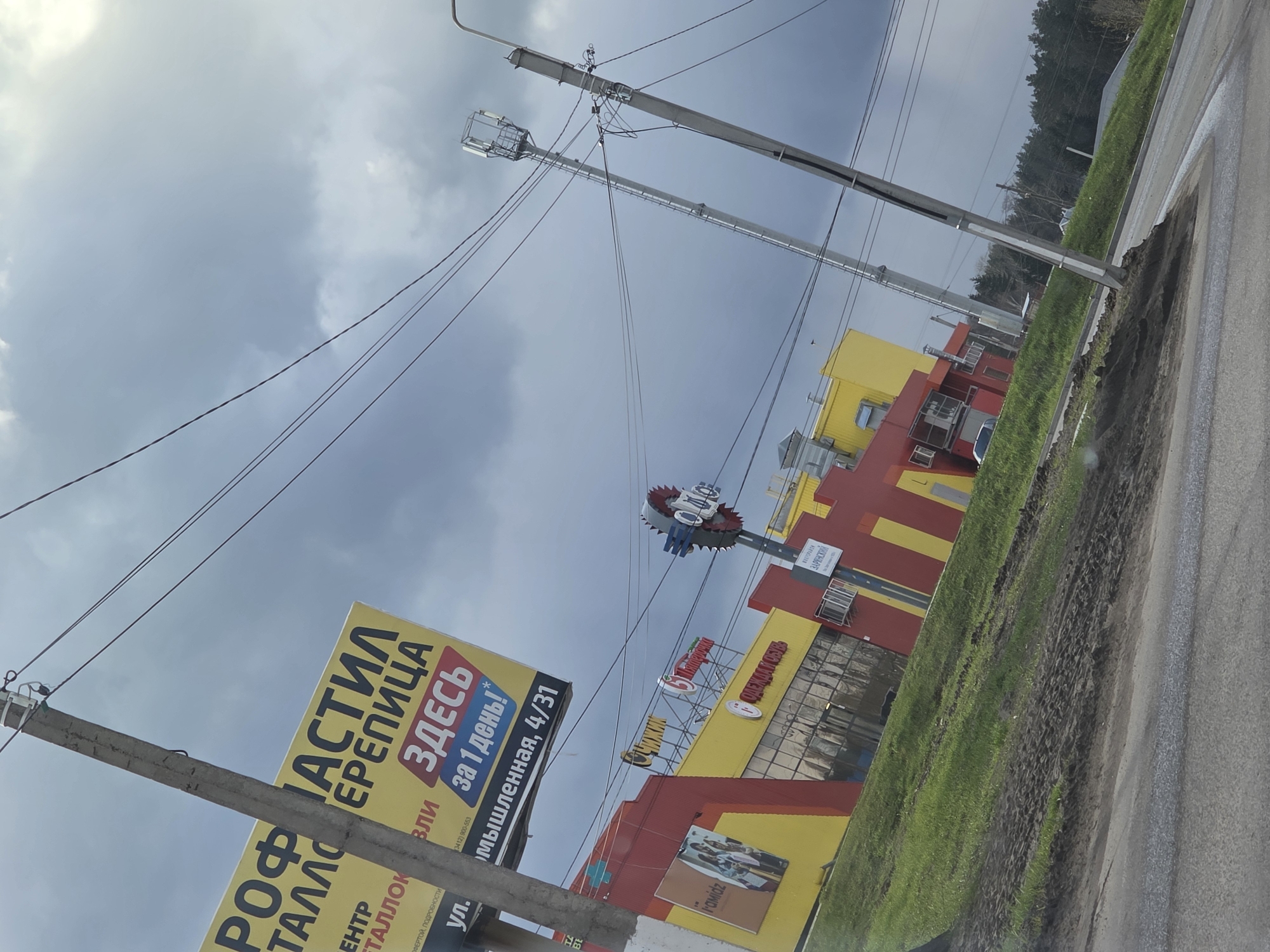 This rotated image captures a street scene in Chaikovsky, Russia, featuring a commercial area under an overcast sky. On the left, a large yellow billboard advertises "ПРОФНАСТИЛ ЕРЕПИЦА" (Corrugated steel, Tile) and "ЦЕНТР МЕТАЛЛОК" (Metal Center), promising service "ЗДЕСЬ за 1 день!" (HERE in 1 day!). It also displays a phone number (3412) 900-553 and an address: "Промышленная, 4/31" (Promyshlennaya St., 4/31).
Multiple utility poles with numerous overhead wires stretch across the frame, indicating an urban or semi-urban setting. To the right, a series of one-story commercial buildings, painted in bright yellow and red, line the roadside. Signs on these buildings identify various establishments, including "Пятерочка" (Pyaterochka), a prominent Russian supermarket chain, "СЧИЖИК" (Chizhik), "ОДЕЖДА И ОБУВЬ" (Clothing and Footwear), and "ЗАРИНСКИЙ" (Zarinskiy). One building features a distinctive red and blue gear-shaped sign on its roof.
The foreground shows a paved road and a grassy verge with patches of mud. In the background, a dense line of trees suggests a forest or wooded area beyond the commercial strip. The sky is largely grey with soft clouds, indicating a cloudy, daytime atmosphere without direct sunlight. No people are visible, and no specific event is taking place, but the scene depicts a typical commercial roadside environment.