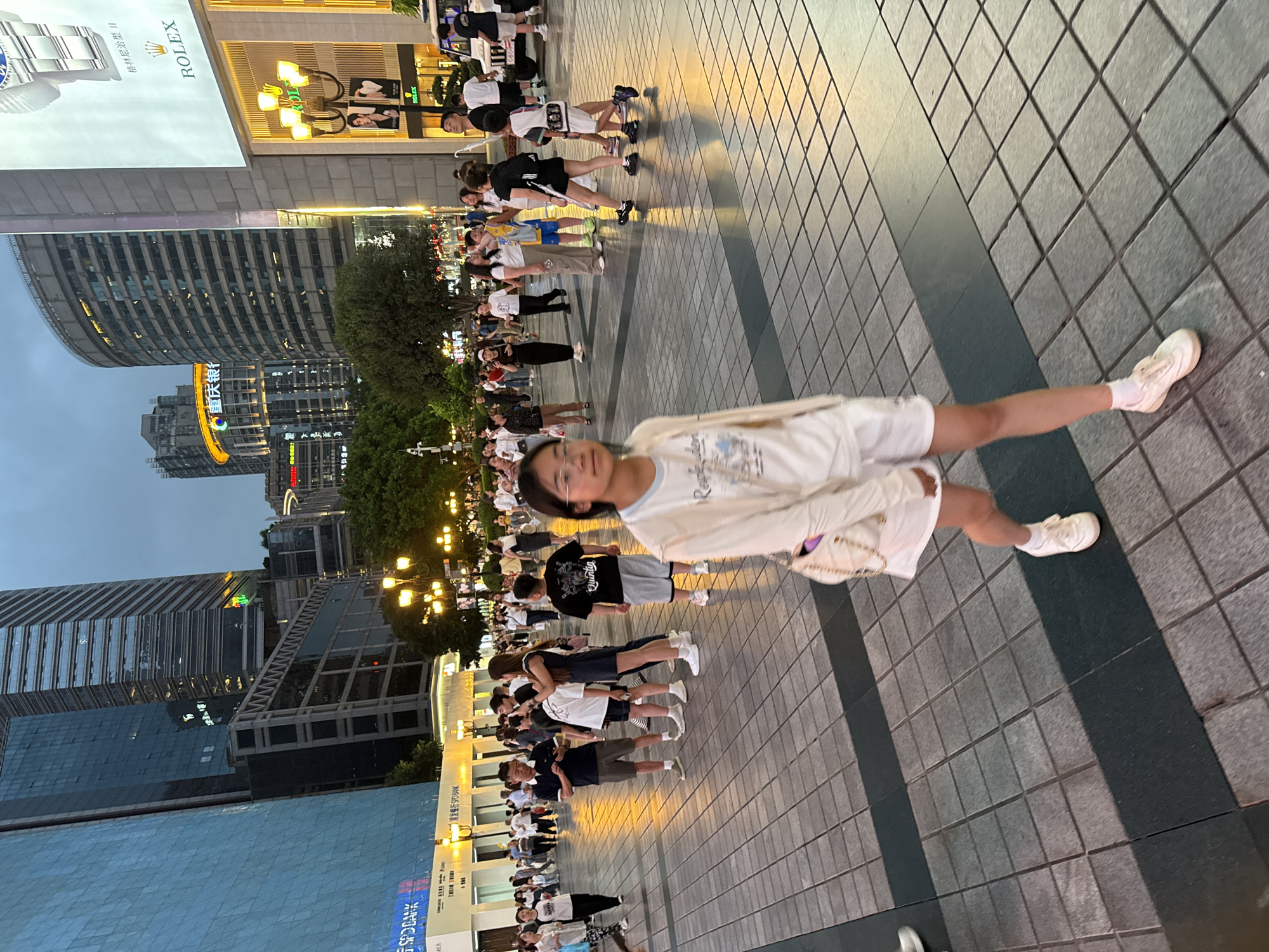 The image depicts a street scene in the Yuzhong District of China, likely in the evening as indicated by the artificial lighting and the twilight sky. Tall, modern buildings, including those with glass facades and illuminated signage, dominate the background, suggesting a commercial or business district.
In the foreground, a young woman with glasses, wearing a light-colored cardigan over a white top and white shorts, is walking towards the viewer. She is carrying a light-colored handbag with a chain strap. The ground is paved with square tiles, some of which are darker, creating a patterned walkway.
Further back, a diverse crowd of people, appearing to be a mix of adults and children, are spread across the plaza. Many are walking, some are standing in small groups, and a few appear to be interacting with their phones. There are several streetlights visible, casting a warm glow on the scene. In the distance, more buildings and signs are discernible, including a prominent Rolex advertisement on a large digital screen. The overall atmosphere is one of a busy urban public space.