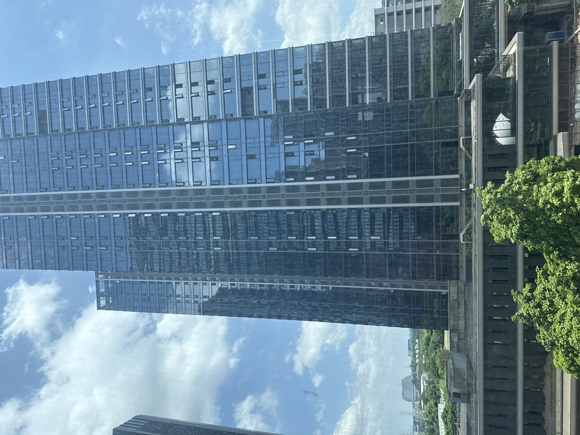 The image depicts a modern cityscape with a prominent glass-facade skyscraper dominating the frame. The skyscraper's reflective surface mirrors the blue sky and scattered white clouds, as well as the surrounding urban environment. In the foreground, lush green trees partially obscure a lower-level building with a dark facade featuring rows of vertical windows. Further in the background, other buildings and structures are visible, including what appears to be a distant, curved building topped with what may be solar panels, and a latticework of electrical transmission towers, suggesting an industrial or infrastructure area. The scene is likely captured from an elevated vantage point, possibly another building or a window, offering a panoramic view of the city. The lighting suggests it is daytime, with bright sunlight. The location is identified as Haizhu District, China.