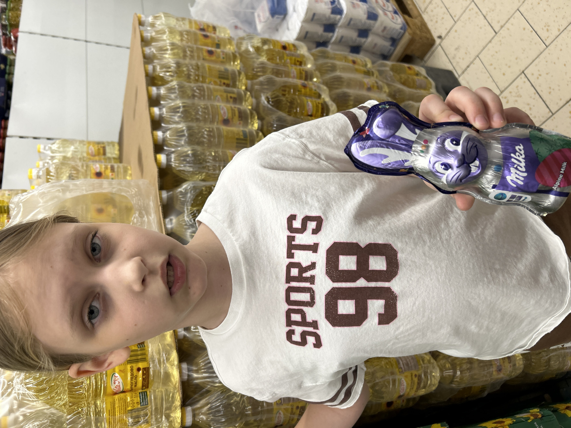 A fair-skinned child is holding a Milka Alpine Milk chocolate bunny in a clear wrapper. The child has blond hair, blue eyes, and is wearing a white t-shirt with "SPORTS 86" printed in brown letters. The background shows multiple rows of plastic bottles filled with yellow oil, possibly sunflower oil. White packages are visible on a pallet behind the child and the oil bottles. The setting appears to be a supermarket aisle. The location is likely Sofia, Bulgaria, given the Cyrillic writing on the oil bottles (Vita d'Or МАСЛО).