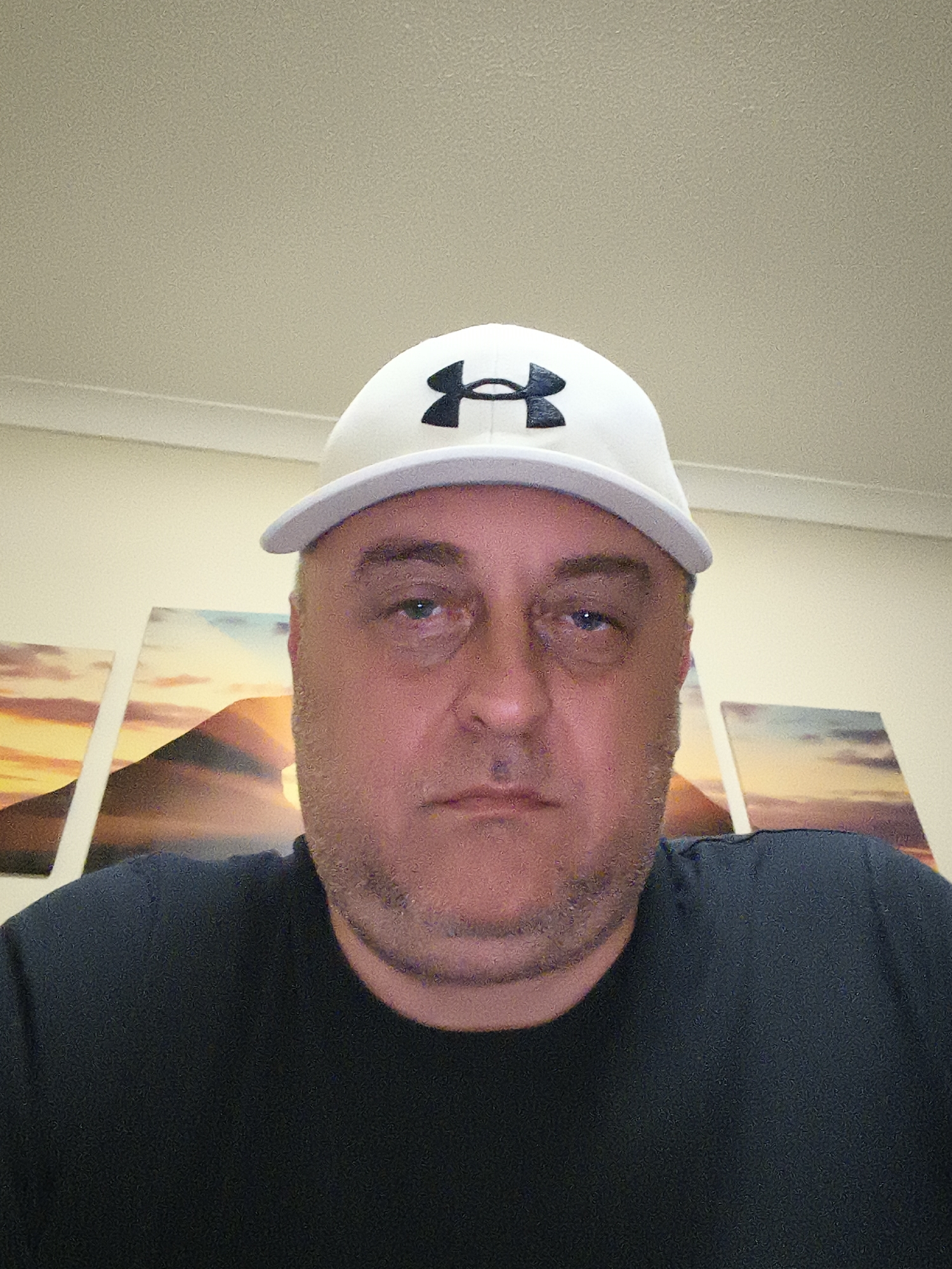 The image shows a man looking directly at the camera. He is fair-skinned, appears to be middle-aged, and has a short beard or stubble. He wears a white baseball cap with a black brim and a prominent black Under Armour logo on the front. He is also wearing a dark, possibly black or navy blue, crew-neck t-shirt. The man's expression is neutral, with his eyes open and looking forward.
The setting appears to be indoors, against a light-colored wall. Behind the man, several horizontal art prints are visible, depicting abstract landscape or sunset scenes with warm orange, yellow, and blue hues, possibly showing mountains or hills. The lighting is consistent with an indoor environment. The image exhibits some digital noise or grain.
**Visible text:** The Under Armour (UA) logo on the cap.