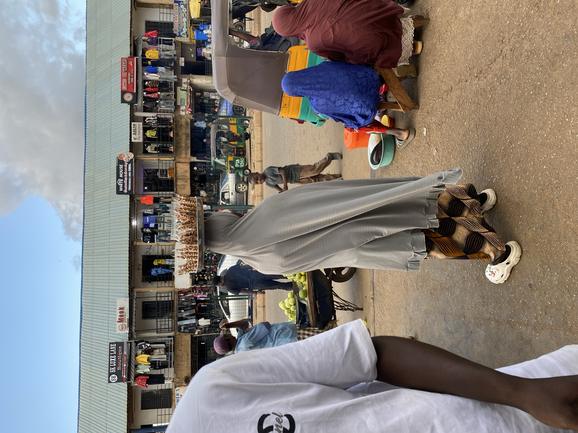 The image captures a busy street scene in Bukuru, Nigeria. A woman with a gray robe walks, balancing a tray of what appears to be snacks on her head. Another woman with a brown robe, a blue headscarf, and a child are seated to the right. A man stands and looks in their direction. In the background, shops line the street with signs such as "SK LUXE LANE Boutique," "WHITE HOUSE," "SAYAD FIT WEARS," "9 SALIM," and "JUDEMORE." Some businesses are displaying clothing items. There are several tricycle vehicles in the background. A person in a white shirt is seated in the foreground.
