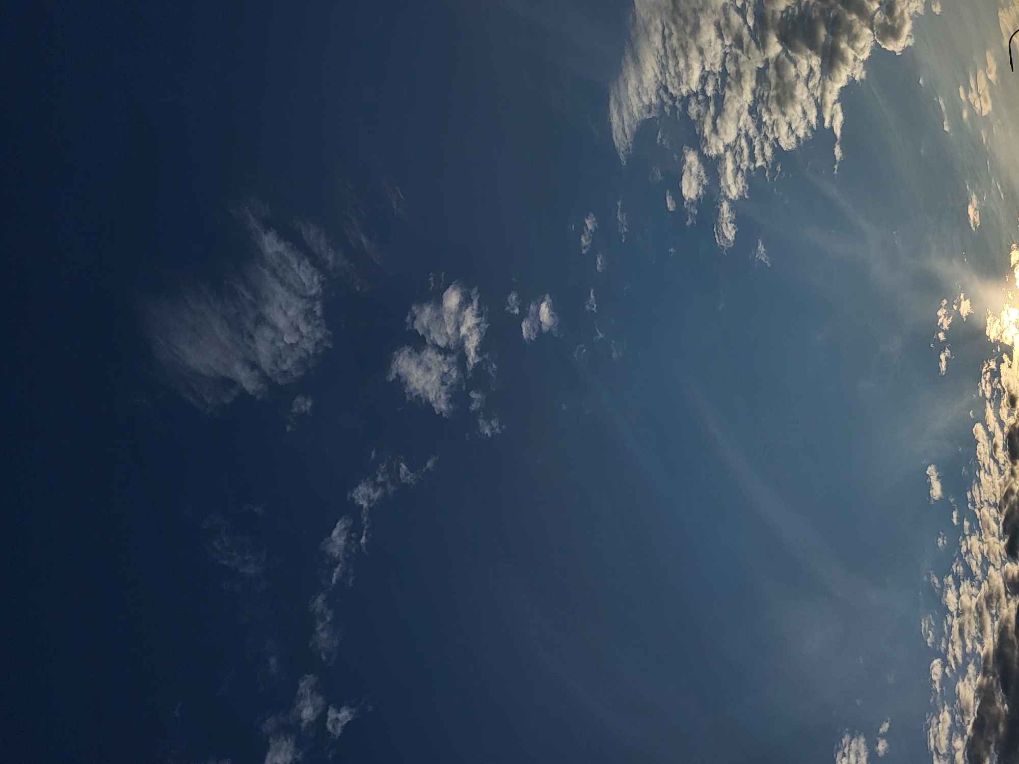 The image depicts a clear sky with scattered clouds above a horizon line. The sky is a deep blue, transitioning to lighter shades towards the bottom. Wispy, thin clouds are visible in the upper portion of the image, while more dense, cumulus-like clouds are present near the horizon. A bright light source, likely the sun, is partially obscured by clouds at the bottom right, casting rays of light upwards. There are no visible people, subjects, or specific objects that would allow for the identification of the city of Jelgava, Latvia, or any particular location within it. The scene is solely focused on the sky and cloud formations.