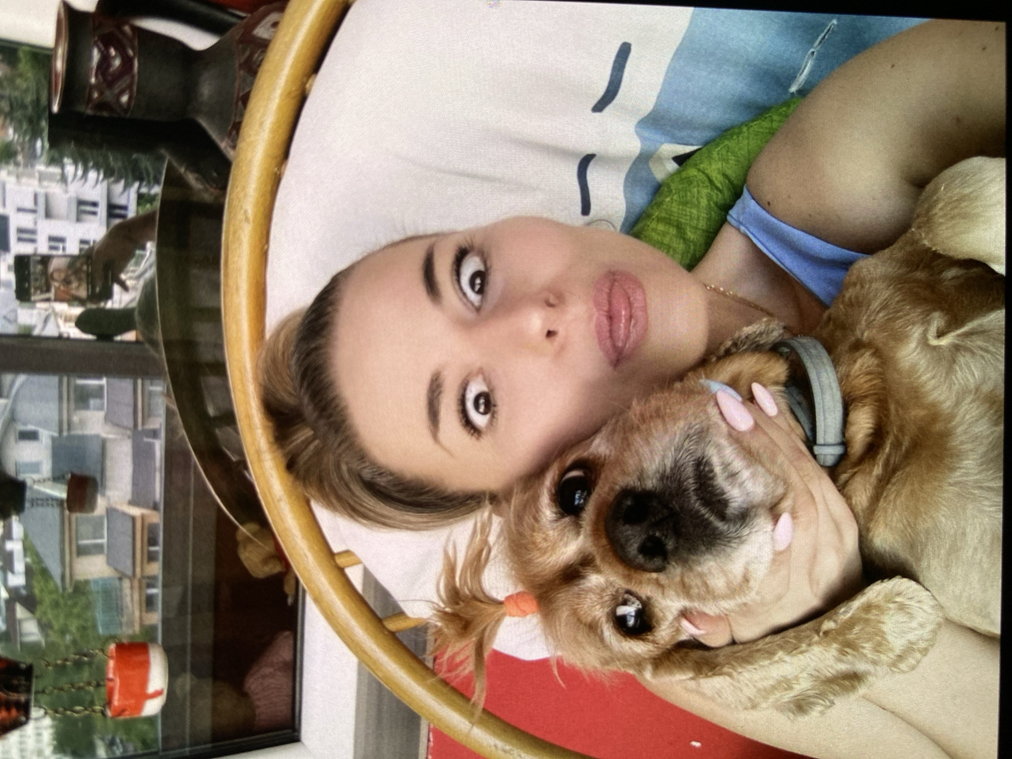 The image shows a young woman and a small dog posing for a photo indoors. The woman, with light brown hair and prominent eyes, is positioned at the center, looking directly at the camera with a playful expression, her lips slightly parted. She wears a light blue top and a delicate gold necklace, and her nails are painted pink.
Cuddled next to her is a small dog with light brown, fluffy fur and dark eyes and nose. The dog wears a grey collar, and a small orange hair tie adorns some fur on its ear. The woman's hand, with pink manicured nails, gently cups the dog's head.
They appear to be resting on a bed or couch, with white bedding featuring blue stripes and a green fabric visible. A curved, light wooden structure, possibly part of a bed frame or chair, is visible on the left.
Through a large window in the background, a daytime scene of Bankya, Bulgaria is visible, showing apartment buildings with balconies and surrounding greenery. Reflections in the window reveal interior elements such as a dark patterned vase and a hanging decoration with red and white components and chains. A faint reflection of a person holding a smartphone suggests the image might be a selfie or taken through a reflection. The overall atmosphere conveys affection and a relaxed, casual moment between the woman and her dog. No text is visible in the image.