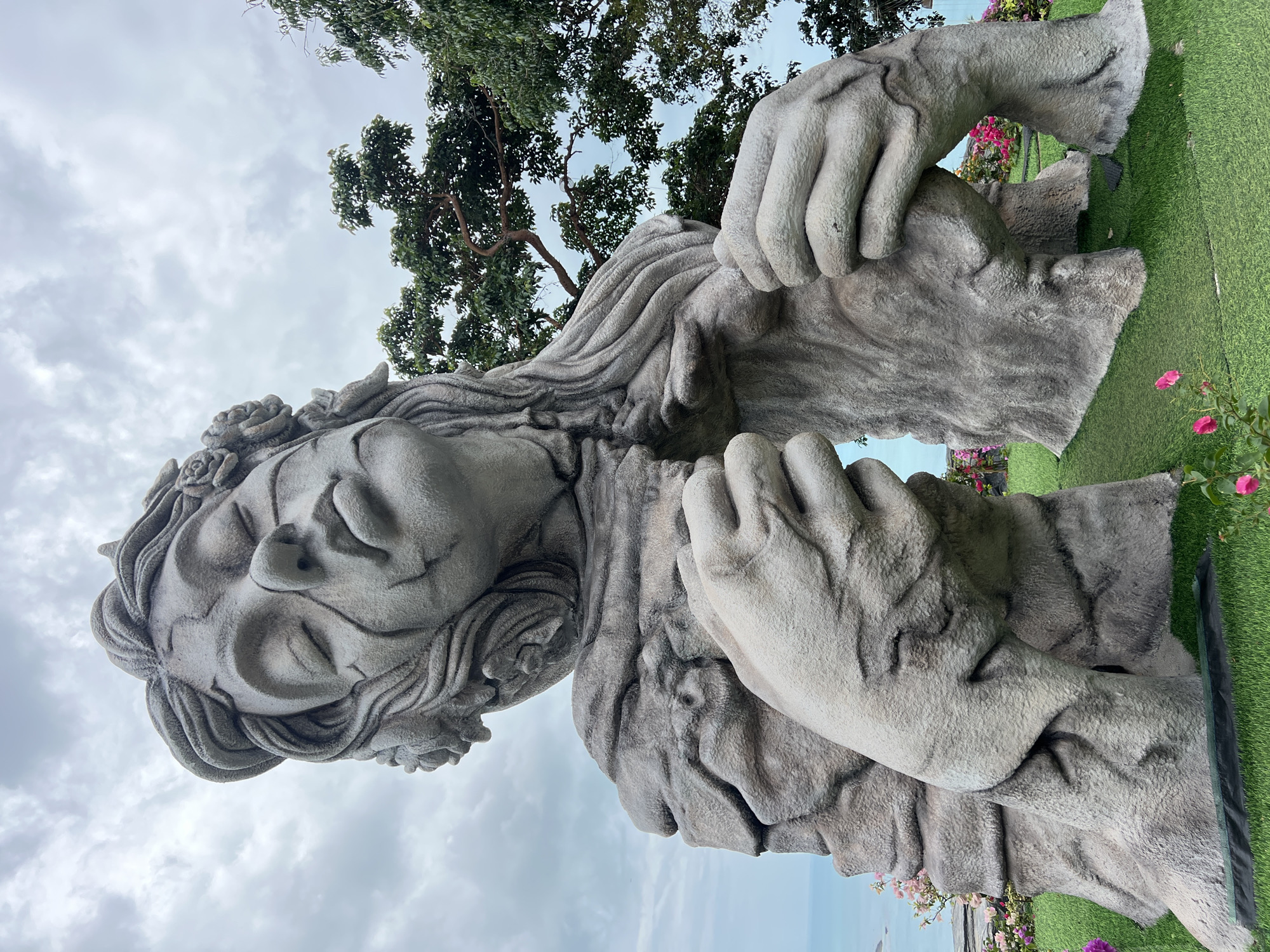 A low-angle, vertical photograph depicts a stone statue of a woman with her eyes closed, holding a textured object, possibly a stone or piece of wood. The statue is set against a backdrop of a cloudy sky and tree branches. The ground features artificial green turf and floral arrangements. The location is the city of Unknown, Unknown.