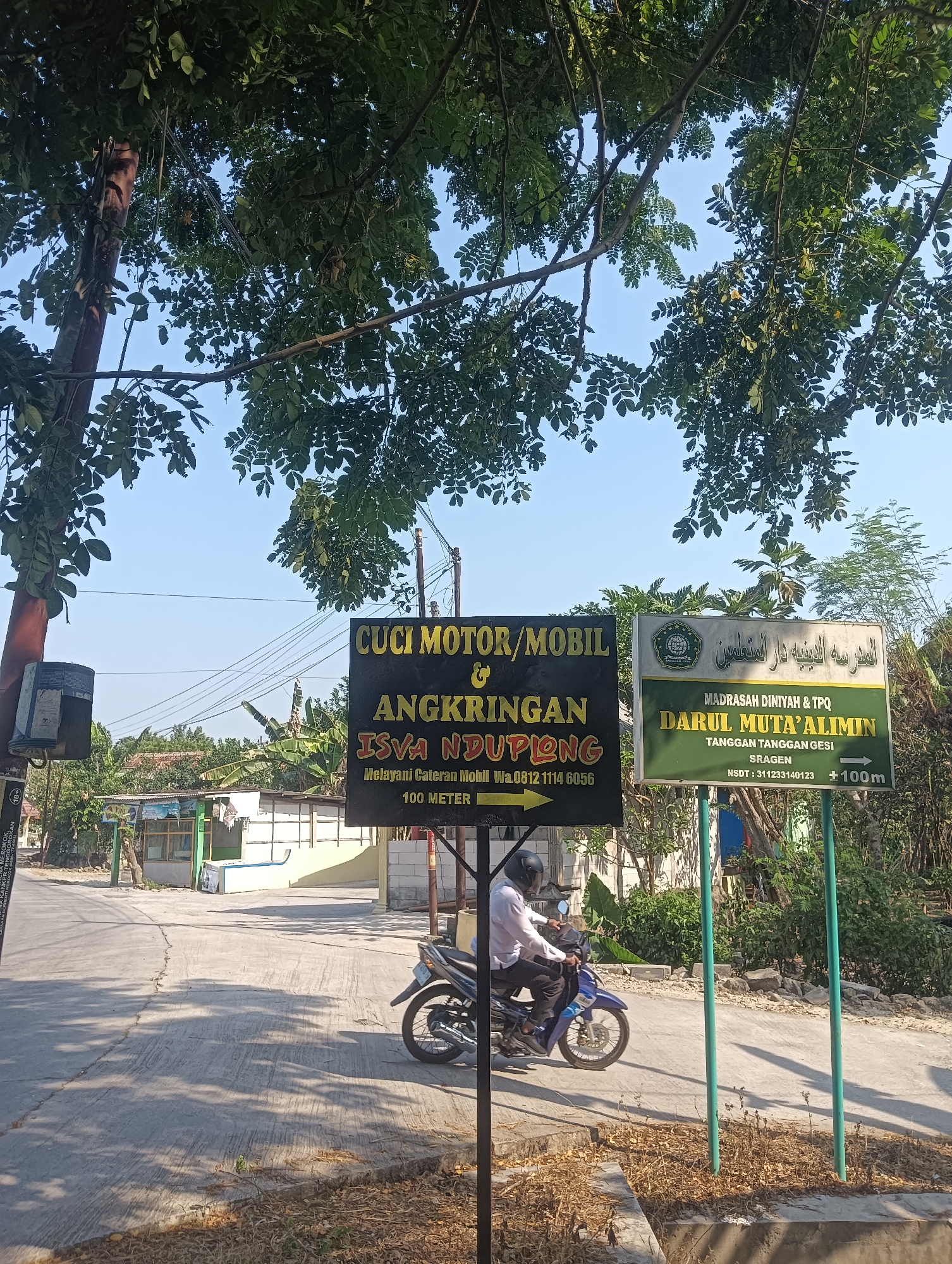 The image depicts a street scene in Tanggan, Indonesia, with two prominent signs. The sign on the left, with a black background and yellow lettering, advertises a car and motorcycle wash and a place called "ANGKRINGAN ISVA NDUPIONG," offering car wash services for "100 METER." Below this, there's a phone number: "0812 1114 6056." The sign on the right, with a green background and yellow lettering, displays "MADRASAH DINIYAH & TPQ DARUL MUTÄ'ALIMIN" (Darul Muta'Alimin Islamic Religious School and Quran Study Center) and "TANGGAN TANGGAN DESI SRAGEN." It also shows a National Unique School Number (NSDT) "311233140123" and a distance of "+100m."
In the foreground, a person wearing a white shirt and black pants, with a black helmet, is riding a blue motorcycle on a paved road. The road has a visible crack running through it. Sunlight casts shadows of trees onto the road. In the background, there are several buildings, including what appears to be a small shop or stall with a green roof, and other structures that suggest a residential or commercial area. Lush green trees and foliage are visible, with some power lines strung across the sky. The overall environment is rural or semi-urban, under a clear blue sky.
