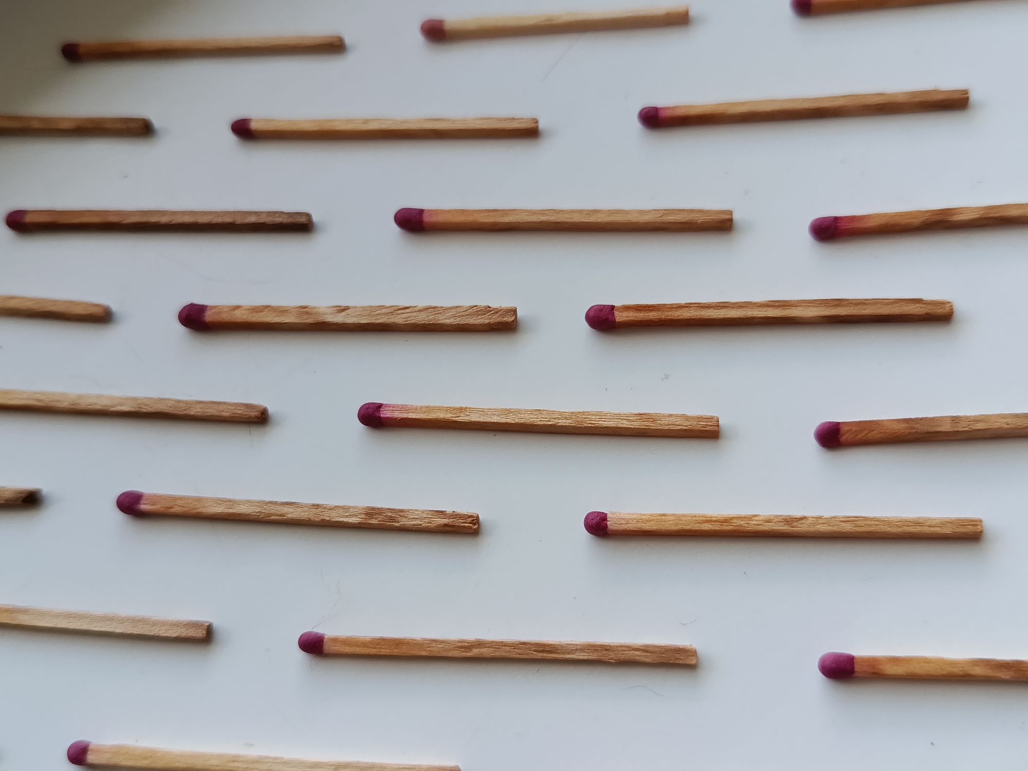An overhead shot displays a white surface with numerous wooden matches scattered across it. The matches are arranged in a somewhat organized pattern, with some rows appearing more uniform than others. Each match has a red tip, indicating it is a safety match. The lighting is neutral, and the focus is sharp, highlighting the texture of the wood and the color of the match heads. There are no people, animals, or other discernible objects in the image. The background is entirely the white surface.