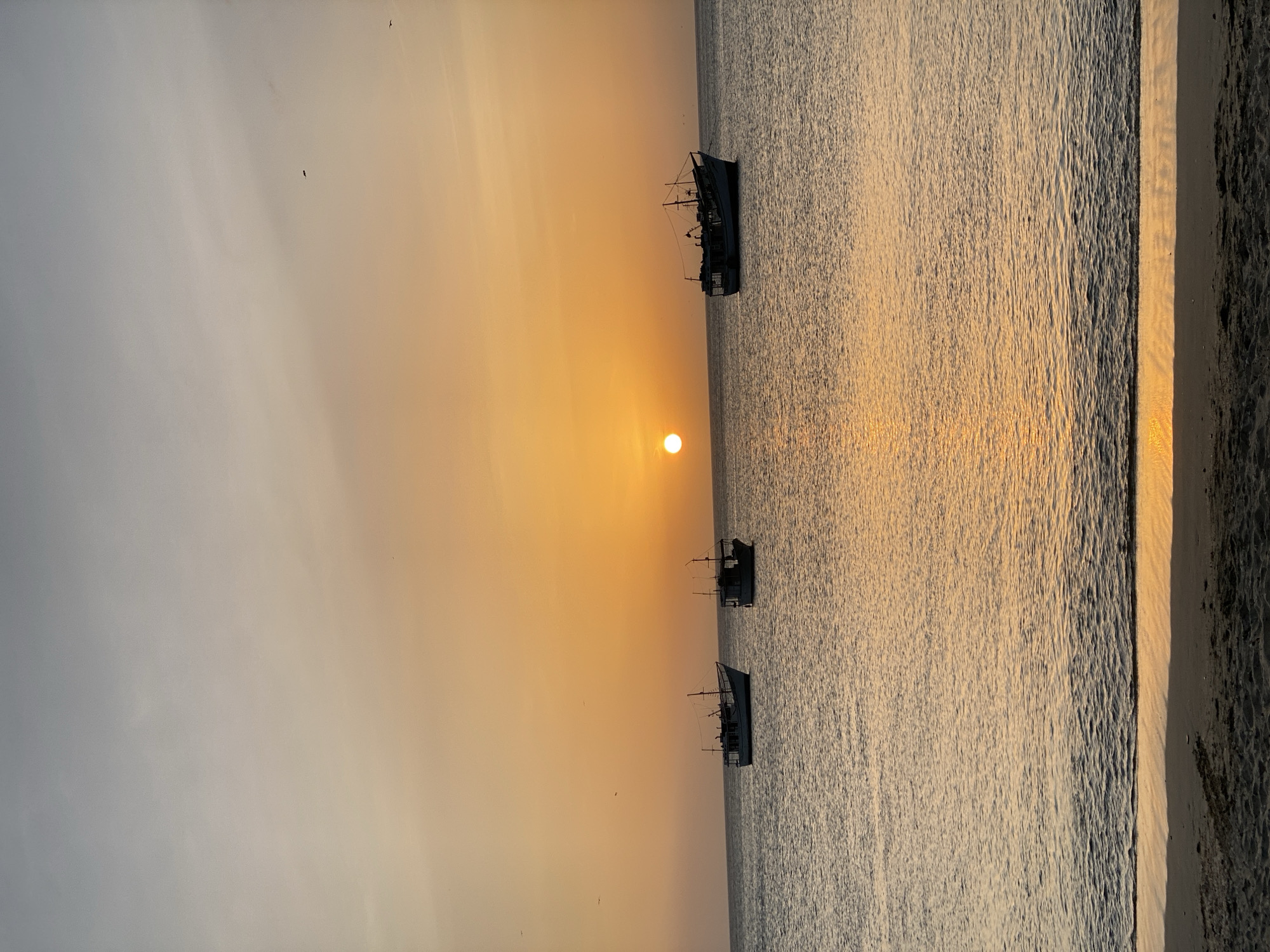 The image captures a serene sunset scene at the beach in Juan Griego, Venezuela. The sun is positioned near the horizon, casting a golden glow across the water and sky. Three fishing boats are silhouetted against the setting sun, floating in the calm ocean. The shoreline is visible on the right side of the image, with sand transitioning from bright reflections to darker shades. Several birds are flying in the sky, adding to the scene's tranquility.