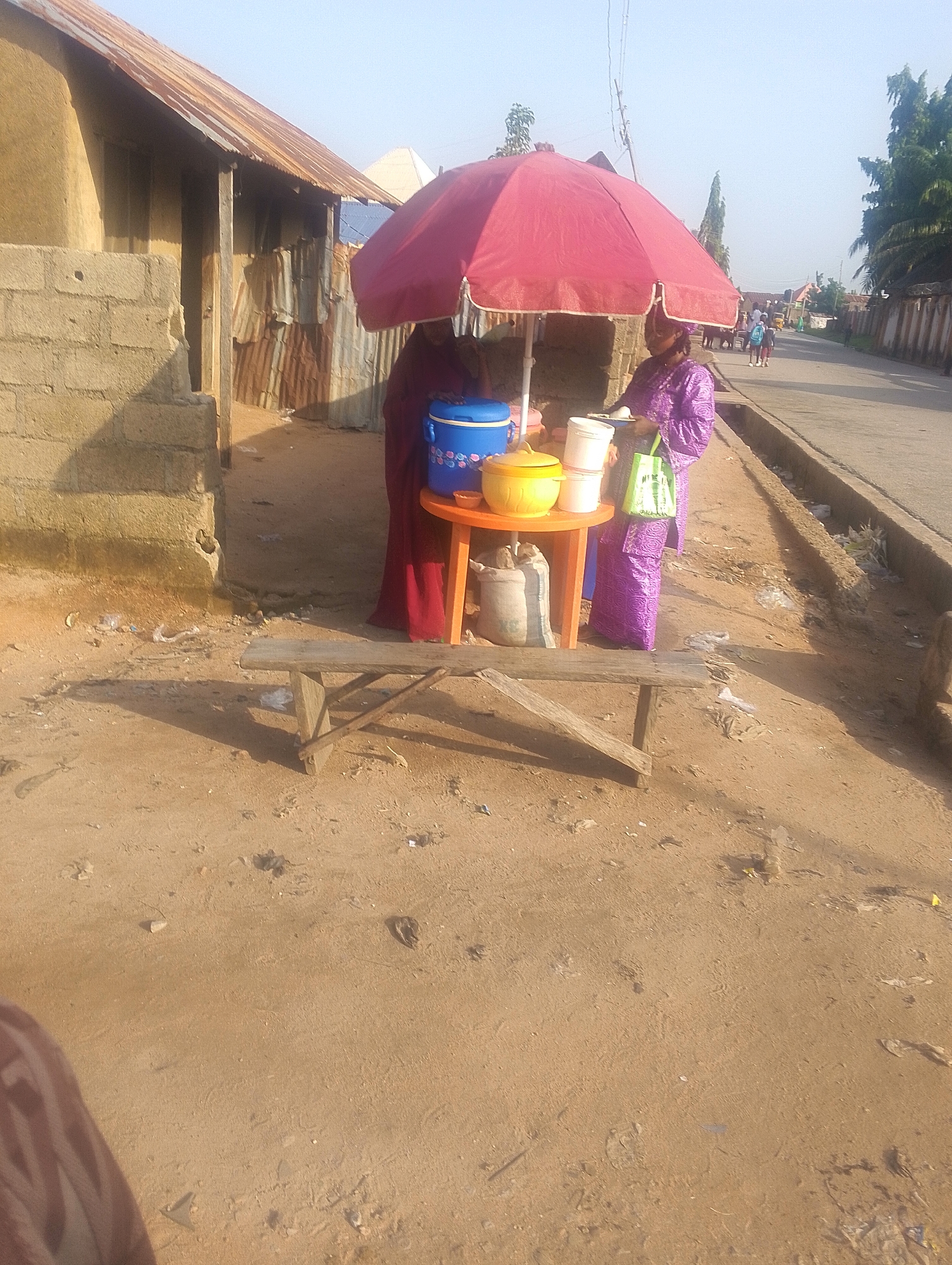 In the city of Jalingo, Nigeria, a roadside vendor is captured under a large red umbrella. The vendor, a woman in a purple patterned garment, stands behind a small orange table displaying several white containers and a yellow insulated jug. Beside the table, a blue insulated cooler sits on the ground, and a large sack is propped against the table's leg. A red fabric, possibly a dress or shawl, is draped over a structure to the left. In the background, a weathered building with a corrugated metal roof and concrete block walls is visible, alongside a dusty, unpaved street with a curb and a drainage ditch. The environment appears arid, with scattered debris on the ground. The sky above is clear and bright, suggesting a daytime scene.