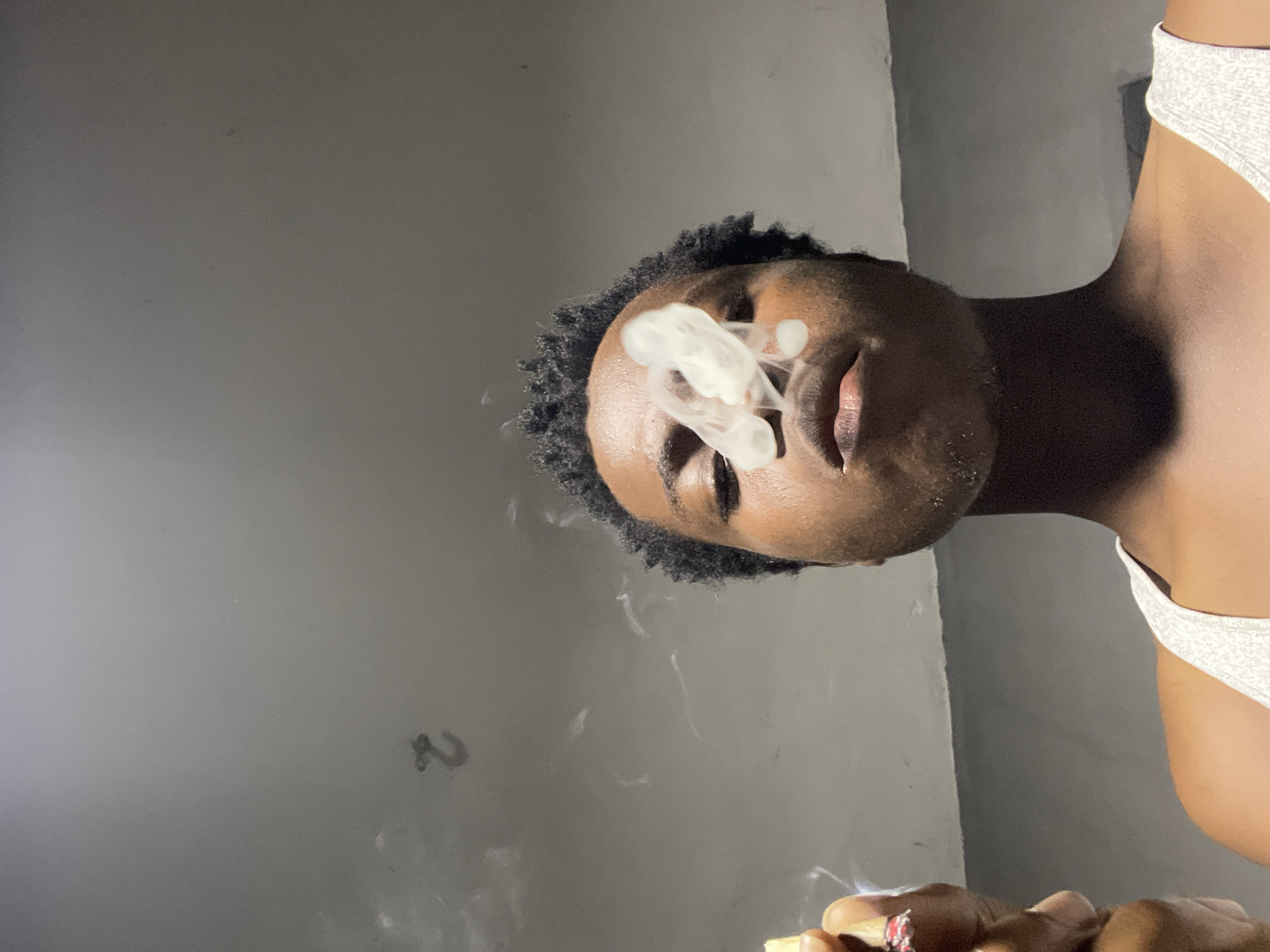 A low-angle shot captures a person exhaling smoke, with a faint "2" marking visible on a grey, textured wall in the background. The person, with dark, textured hair and brown skin, is wearing a white tank top. Their eyes are closed, and smoke billows around their face, obscuring part of their nose and mouth. In their left hand, held near their face, is a lit cigarette. The lighting is dim, creating shadows. The scene appears to be indoors.