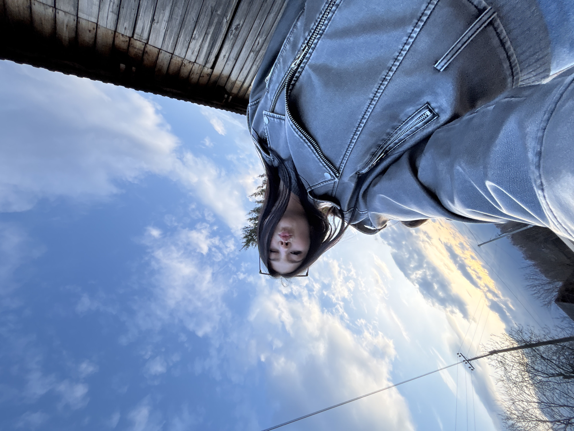 A woman with long dark hair, wearing a gray leather jacket and dark-rimmed glasses, is taking a selfie from a low angle. Above her is a bright blue sky with scattered white clouds. To the left, weathered wooden planks of a building overhang the scene. In the distance, telephone poles and bare tree branches are silhouetted against a yellow-tinged sky. The location is Фоки, Russia.