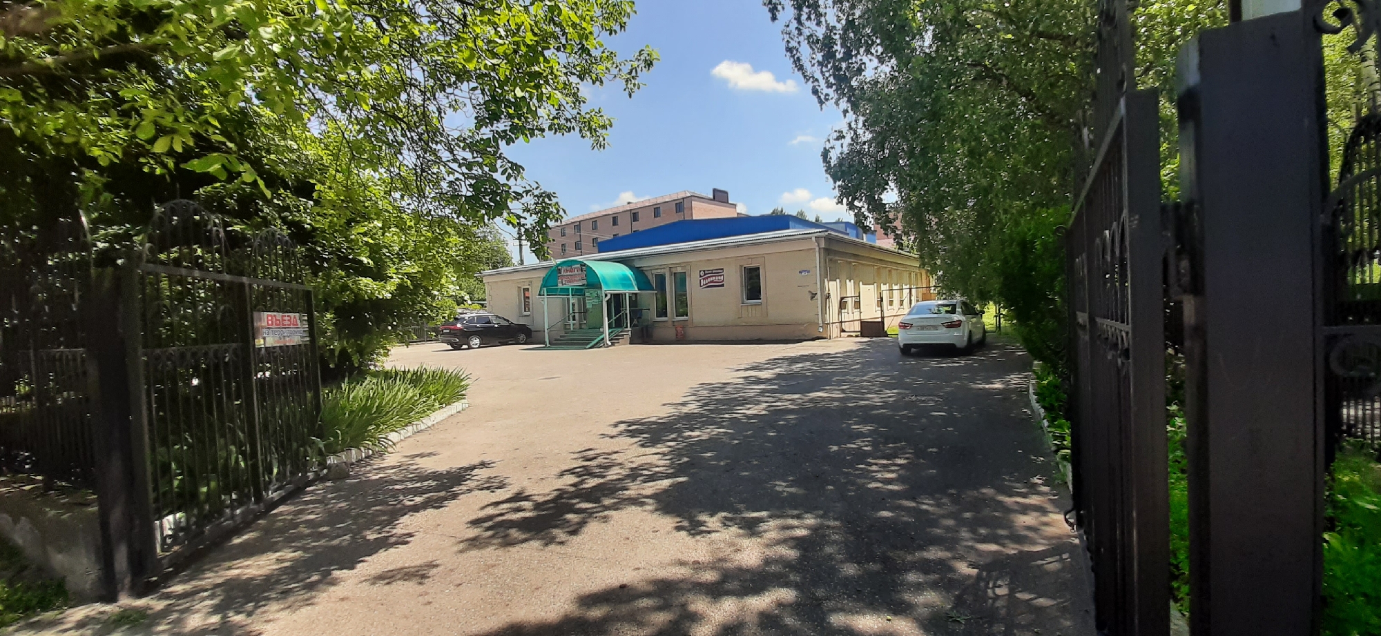 The image shows a street view of a building in Yessentukskaya, Russia, on a sunny day. The building is light-colored with a blue roof and a green awning over the entrance. There are two cars parked in front of the building, one black and one white. A black metal gate with a sign that reads "ВЪЕЗД НА ТЕРРИТОРИЮ" (Entrance to the territory) is visible on the left side of the frame, partially obscured by trees. The area around the building is paved, with trees and greenery surrounding it. In the background, a multi-story building is visible. The overall scene depicts a commercial or administrative building in a semi-urban environment.