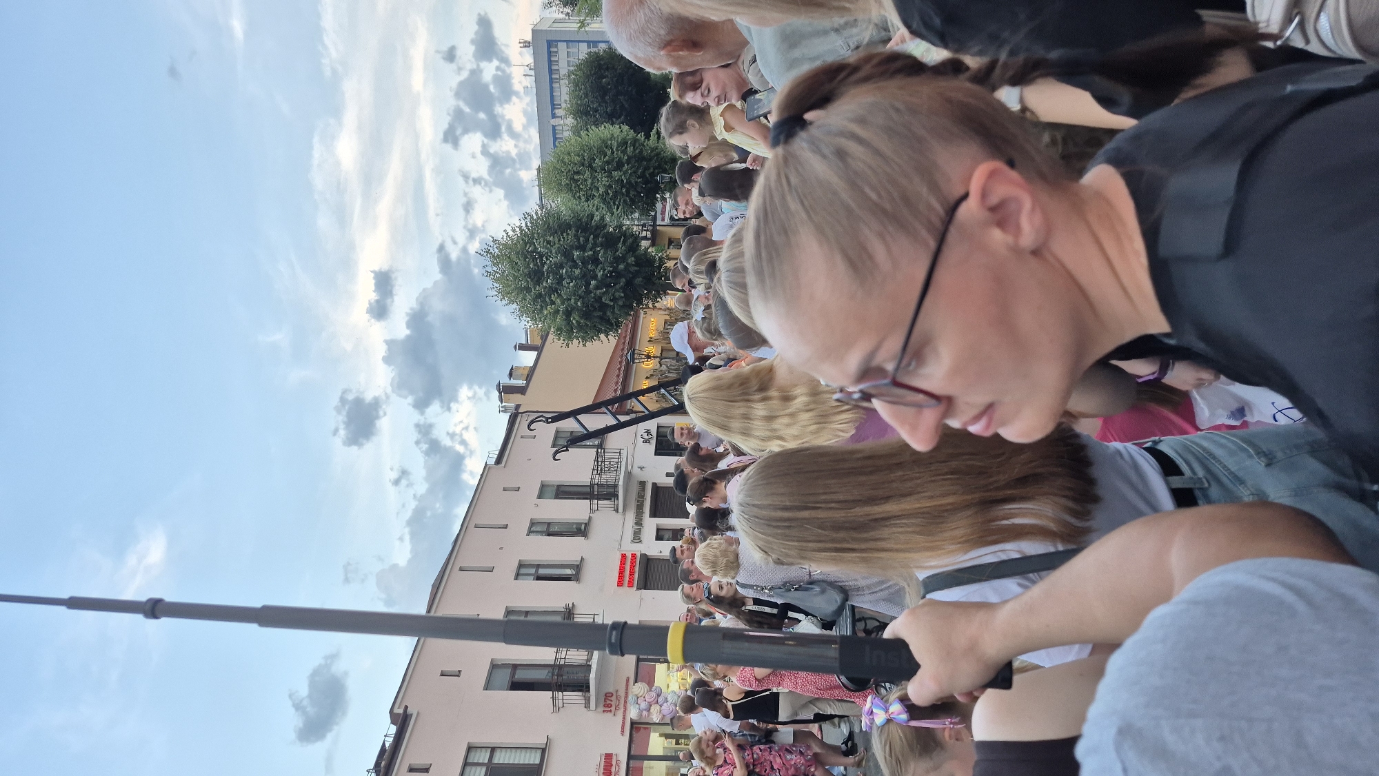 In the city of Brest, Belarus, a crowd of people gathers outdoors on a street lined with buildings. A tall, dark pole, possibly part of a streetlamp or flagpole, stands prominently on the left side of the frame. In the foreground, a woman wearing glasses and a black shirt is looking upwards and to the right, her hair pulled back in a ponytail. Behind her, other individuals are visible, some with their backs to the camera. The background shows a street with shops and buildings, a ladder leaning against one of the structures, and trees. The sky above is partially cloudy, suggesting it may be late afternoon or early evening.