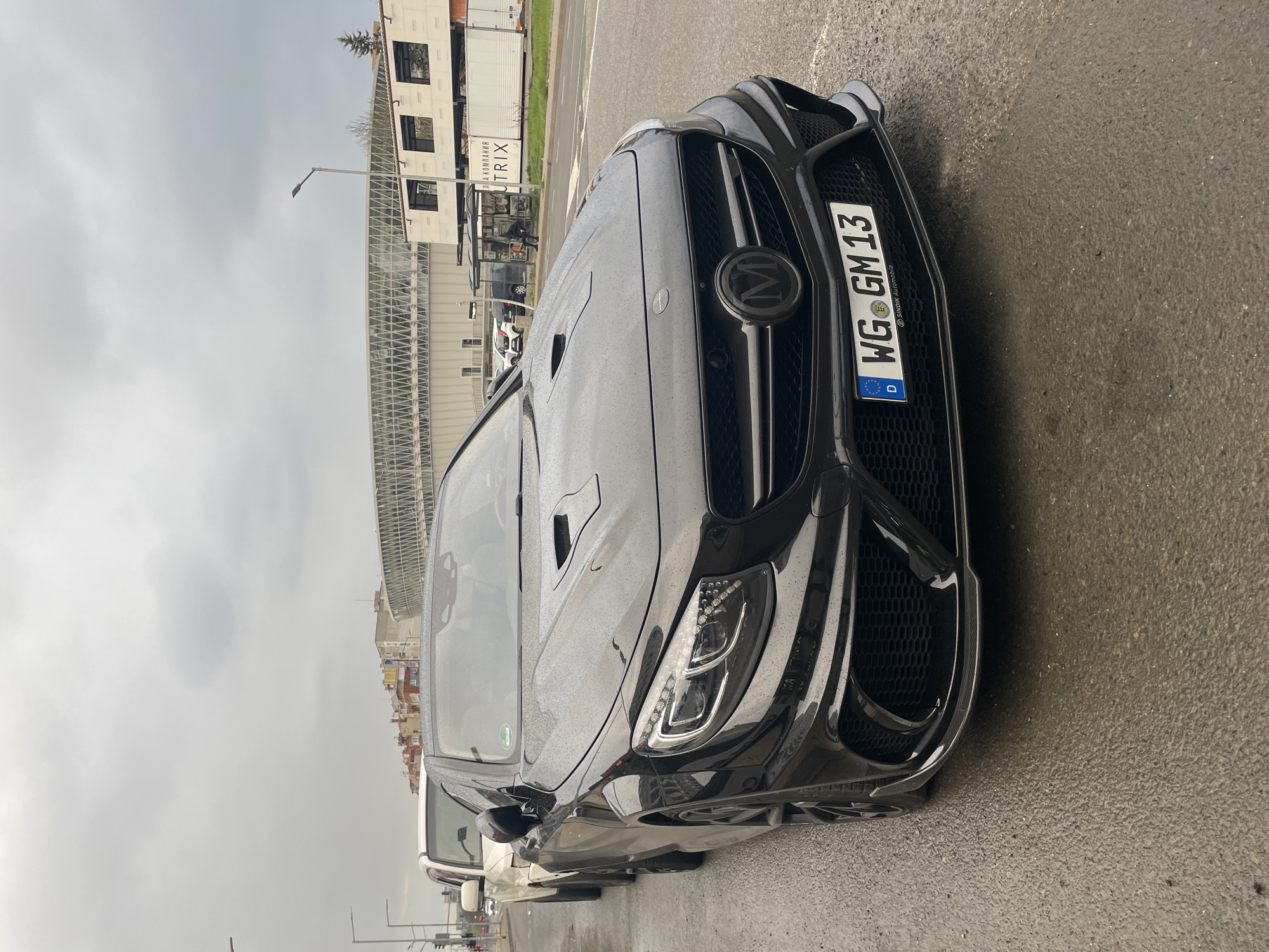 The image shows the front of a black Mansory-modified Mercedes-Benz AMG parked on a street. The car features a custom grille with a blacked-out Mansory emblem, a hood with vents, and a German license plate reading "WG GM 13." Raindrops are visible on the car's surface. In the background are buildings, including one with a glass-covered pedestrian walkway. The location is likely in Burgas, Bulgaria, based on the license plate, which indicates the car is registered in Germany. The scene appears to be taken on a cloudy day. Other vehicles are parked along the street behind the Mansory AMG.