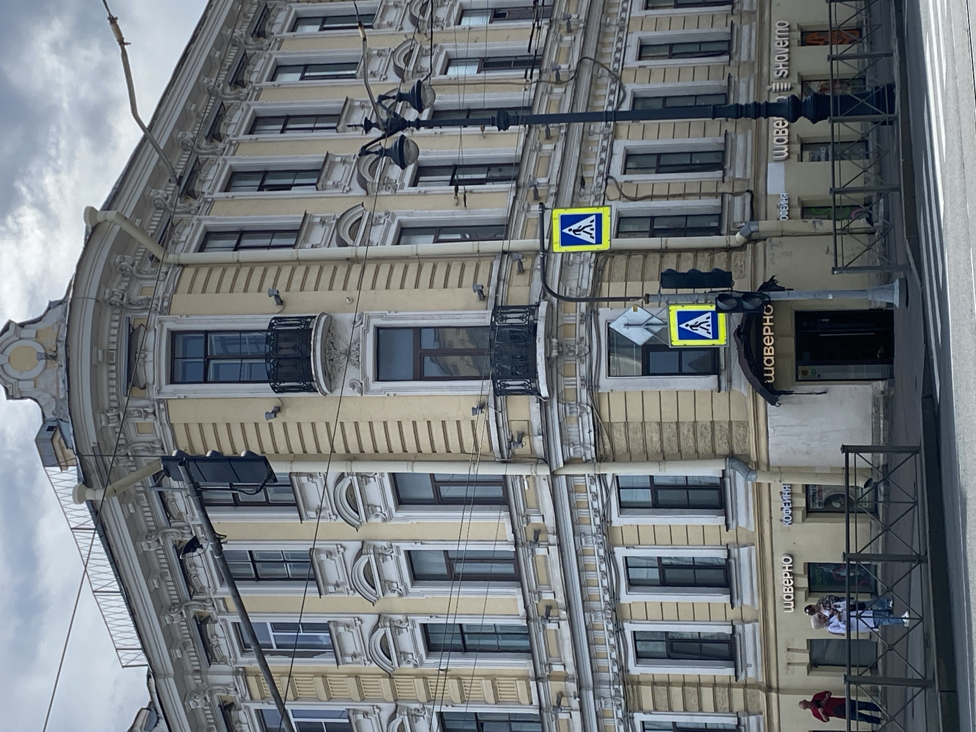 The image depicts a corner view of a light yellow, ornate building in Saint Petersburg, Russia, under a cloudy sky. The building features multiple stories with rows of dark-framed windows, elaborate white decorative moldings, and a rounded corner. A black wrought-iron balcony is visible on the upper level.
In the foreground, a pedestrian crossing is marked on the asphalt road. A dark lamppost with two globe-shaped lights stands near the corner. Two traffic signals are present: one is a standard traffic light, and another is a pedestrian crossing signal showing a walking figure in blue within a yellow square. A smaller blue and white diamond-shaped sign is mounted above the pedestrian signal.
On the sidewalk in front of the building, there are several storefronts with illuminated signage in Cyrillic letters, including "ШАВЕРНО" (Shaverno) and "КОФЕЙНЯ" (Kofeynya - coffee shop). A black metal fence runs along the sidewalk. Three individuals are visible near the fence: one man in a red shirt standing to the left, and a woman in a white headscarf and blue jacket, and another person in a dark shirt and hat to her right.
The overall scene suggests an urban environment, likely a commercial street in Saint Petersburg, characterized by its historical architecture and typical street furniture.