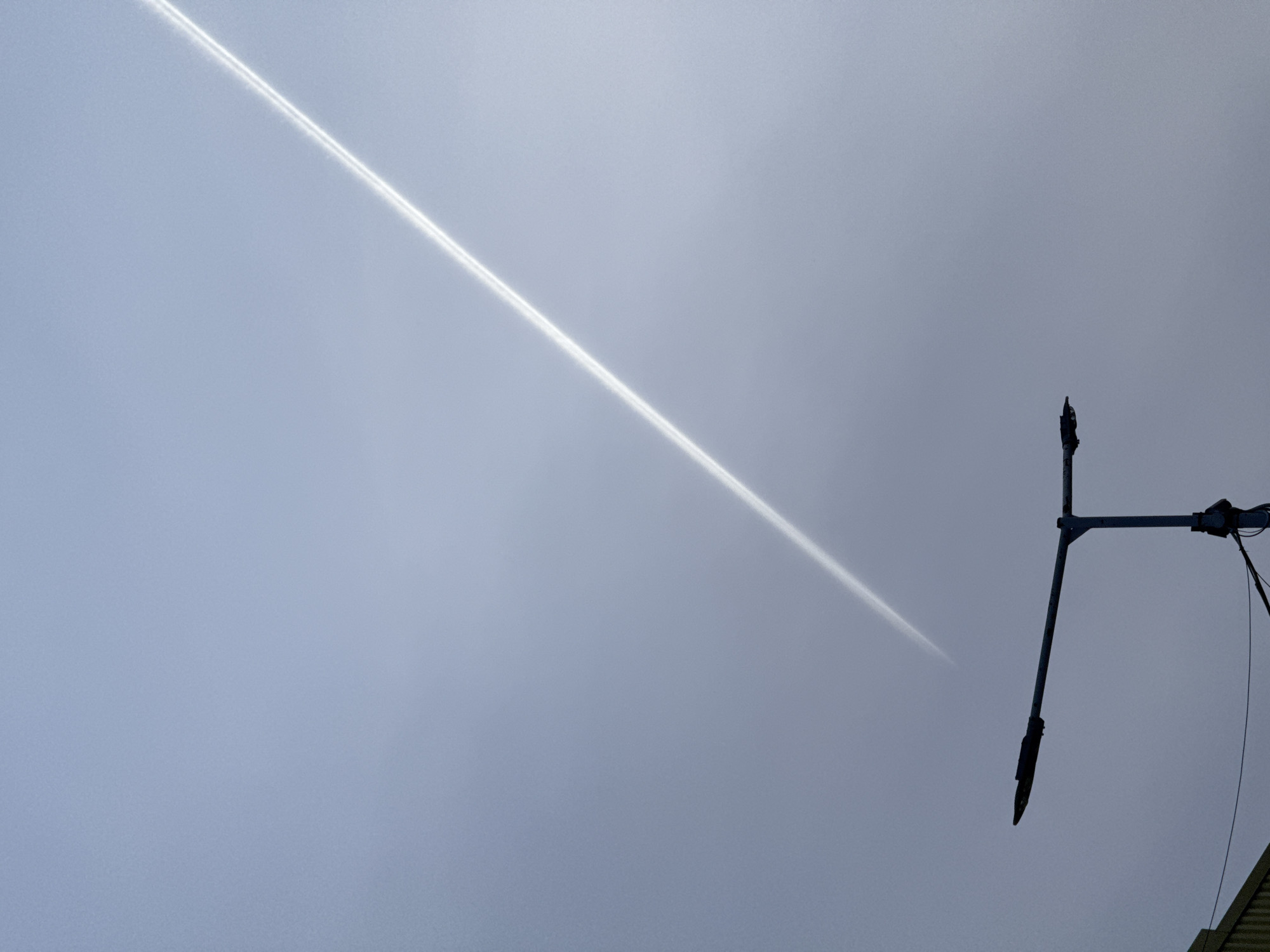 A contrail is visible across a gray sky, partially obscured by clouds. In the lower right corner, a utility pole with equipment is mounted on it. The equipment appears to be black and gray. A section of a roof can be seen below the pole. The sky is overcast and gray.