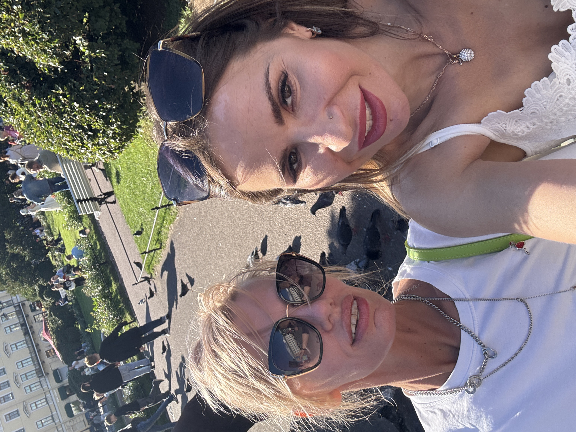 Two women are taking a selfie outdoors on a sunny day. One woman is in the foreground, wearing a white t-shirt and sunglasses. She has blonde hair and is smiling. The other woman, in the background, is wearing a white lace top and sunglasses. She has brown hair and is also smiling. Both women are wearing necklaces. In the background, there are people sitting on a bench, trees, and a building that appears to be in Saint Petersburg, Russia. Pigeons are scattered on the ground around the women. The lighting suggests it is daytime.