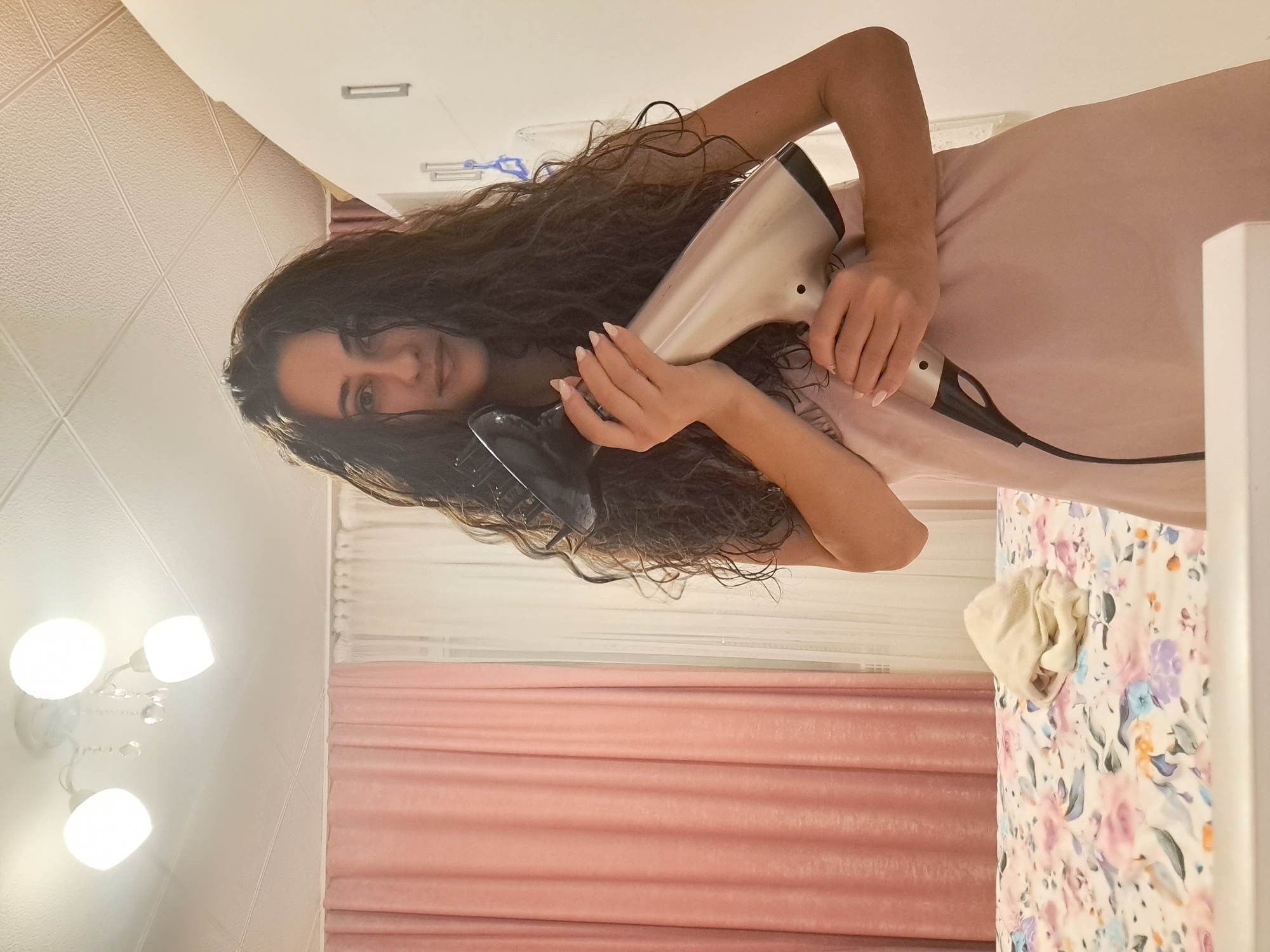 The image depicts a young woman with long, curly dark hair, holding a silver and black hairdryer with a diffuser attachment. She is wearing a pale pink, sleeveless dress. Her fingernails are painted a light, neutral color. She is standing in what appears to be a bedroom. In the background, a bed with floral-patterned bedding is visible. A light-colored cloth is folded on the bed. To the left, pink curtains are drawn, and to the right, a white closet door or wall is present. A ceiling light fixture with three bulbs and decorative crystals hangs overhead. The overall setting suggests an indoor domestic environment. This location is specified as being in the city of Negovan, Bulgaria.