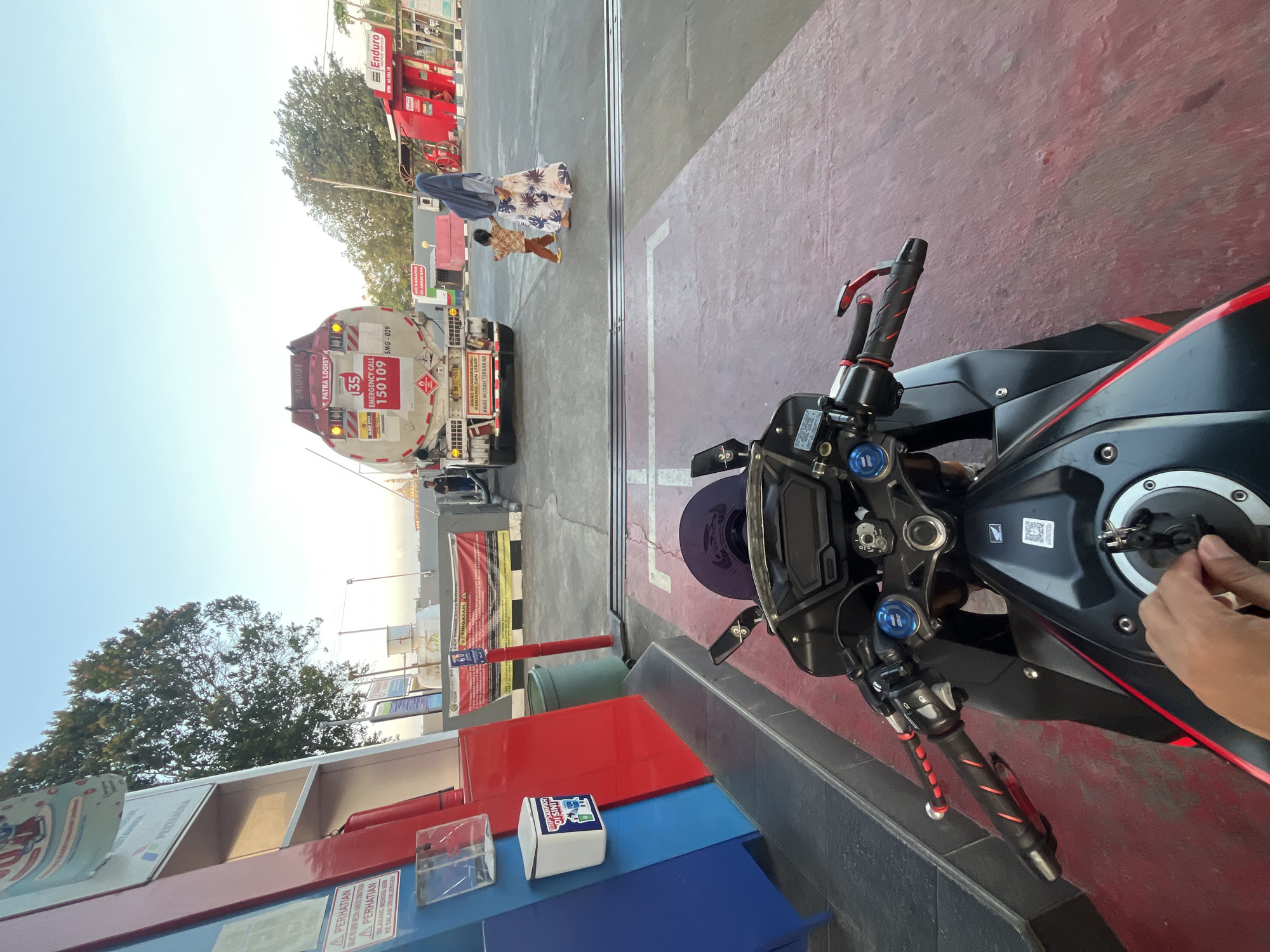 The image depicts a scene at a Pertamina gas station in Pecangaan, Indonesia, possibly at dusk or dawn given the sky's illumination. In the foreground, a motorcycle's handlebars and fuel tank are visible, with a hand reaching towards the ignition. A person's hand is seen inserting a key into the ignition. The gas station features a blue and red structure to the left, possibly part of a pump or service area. Further into the scene, a large white tanker truck with "T. PATRA LOGIST." and identifying numbers "135" and "150109" is parked, suggesting a fuel delivery. A woman in a hijab and a child in a patterned shirt are walking away from the truck towards a red-colored building with "Enduro" signage, likely another service point. The ground is paved and marked with white lines, typical of a gas station forecourt. The background shows trees and a clear sky. The overall environment appears to be an open, outdoor commercial space.