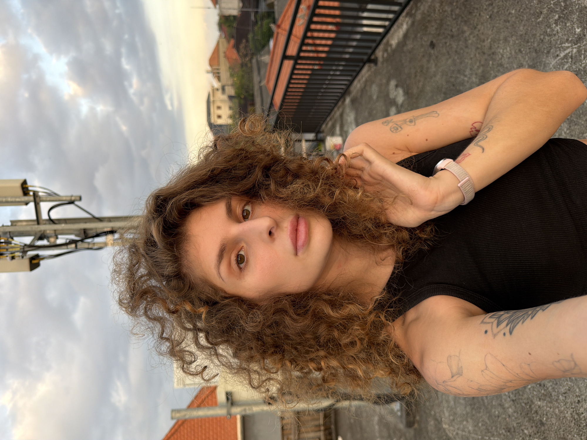 The image is a portrait of a young individual, likely female, with abundant, light brown, curly hair and brown eyes. She looks directly at the camera with a neutral expression, her lips slightly parted. She wears a dark, possibly black, ribbed top. On her left wrist, a light pink or beige fabric smartwatch band is visible. Her arms display several tattoos: a simple outline of a guitar-like shape and a red line on her right forearm, and faint floral outlines and a partial mandala design on her left arm. Her hands are gently clasped near her chest, with one hand lightly touching her hair.
The setting is an outdoor rooftop or elevated terrace, providing a view of an urban landscape in North Kuta, Indonesia. Buildings with characteristic terracotta-colored tiled roofs are seen in the background under a cloudy sky, suggesting the late afternoon or early evening (golden hour) due to the soft, warm lighting. To the far left, a metallic utility structure with cables is partially visible. The person is posing for the camera in a relaxed manner.