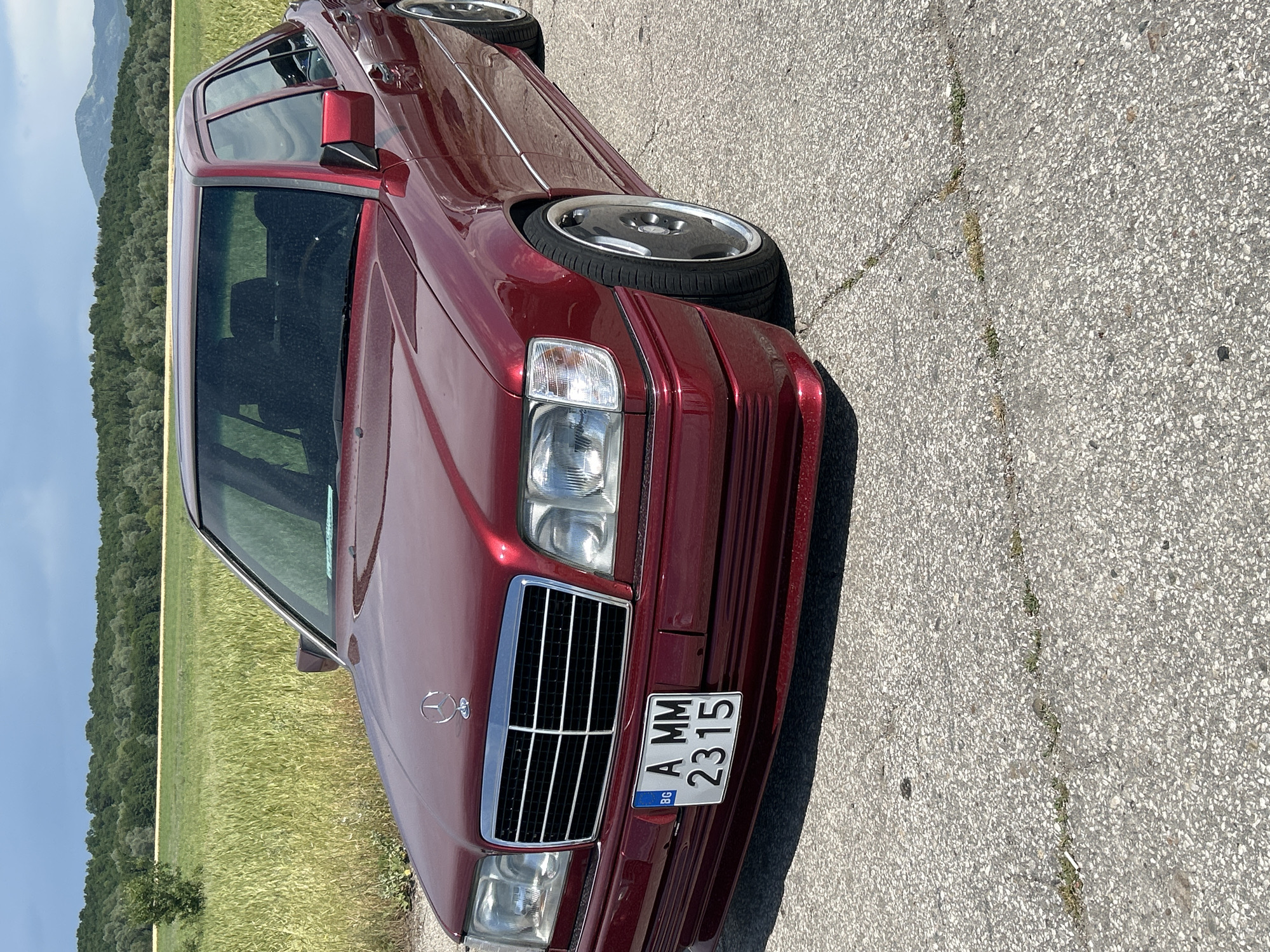 A maroon Mercedes-Benz W124 is parked on an asphalt road with the license plate "BG A MM 2315." The car is positioned at an angle, showcasing its front and side. The background reveals a field of tall grass and a tree line, suggesting a rural or semi-rural setting. There is a mountain range in the distance under a clear sky. The car is the primary object of interest. No people are visible in the image. The location may be in the vicinity of Dalbok Izvor, Bulgaria, given the license plate's "BG" code. The condition of the road appears cracked and worn.