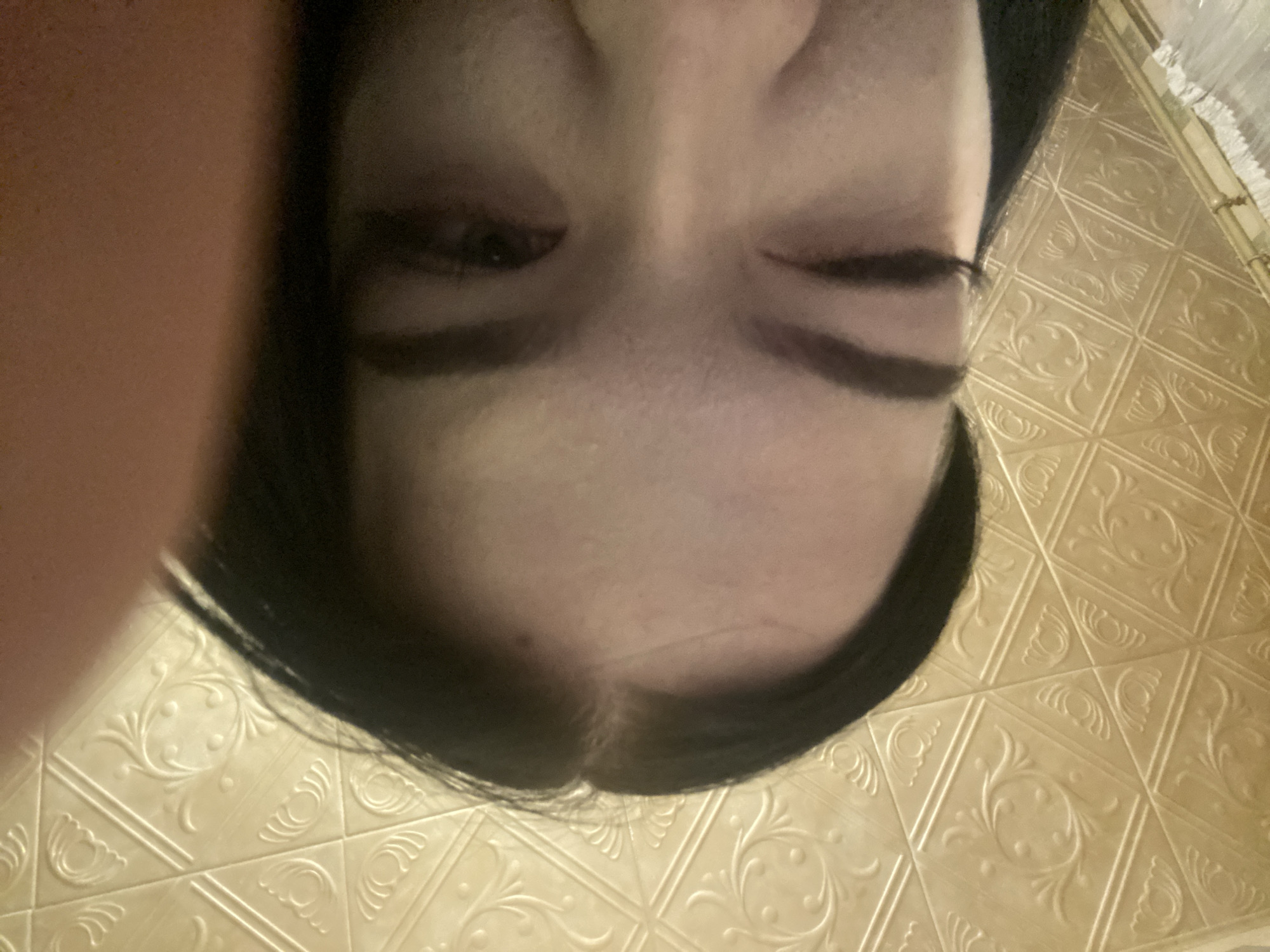 An overhead close-up shot depicts a person's face, primarily focusing on the forehead, nose, and closed eyes with prominent dark eyebrows. The person has dark hair, and their face is relatively pale. The shot is taken at an unusual angle, with the person's face tilted toward the camera.
The background consists of a tiled surface with an ornate, embossed pattern. The tiles appear to be a light, yellowish-beige color. The location is not possible to identify based on the given information.